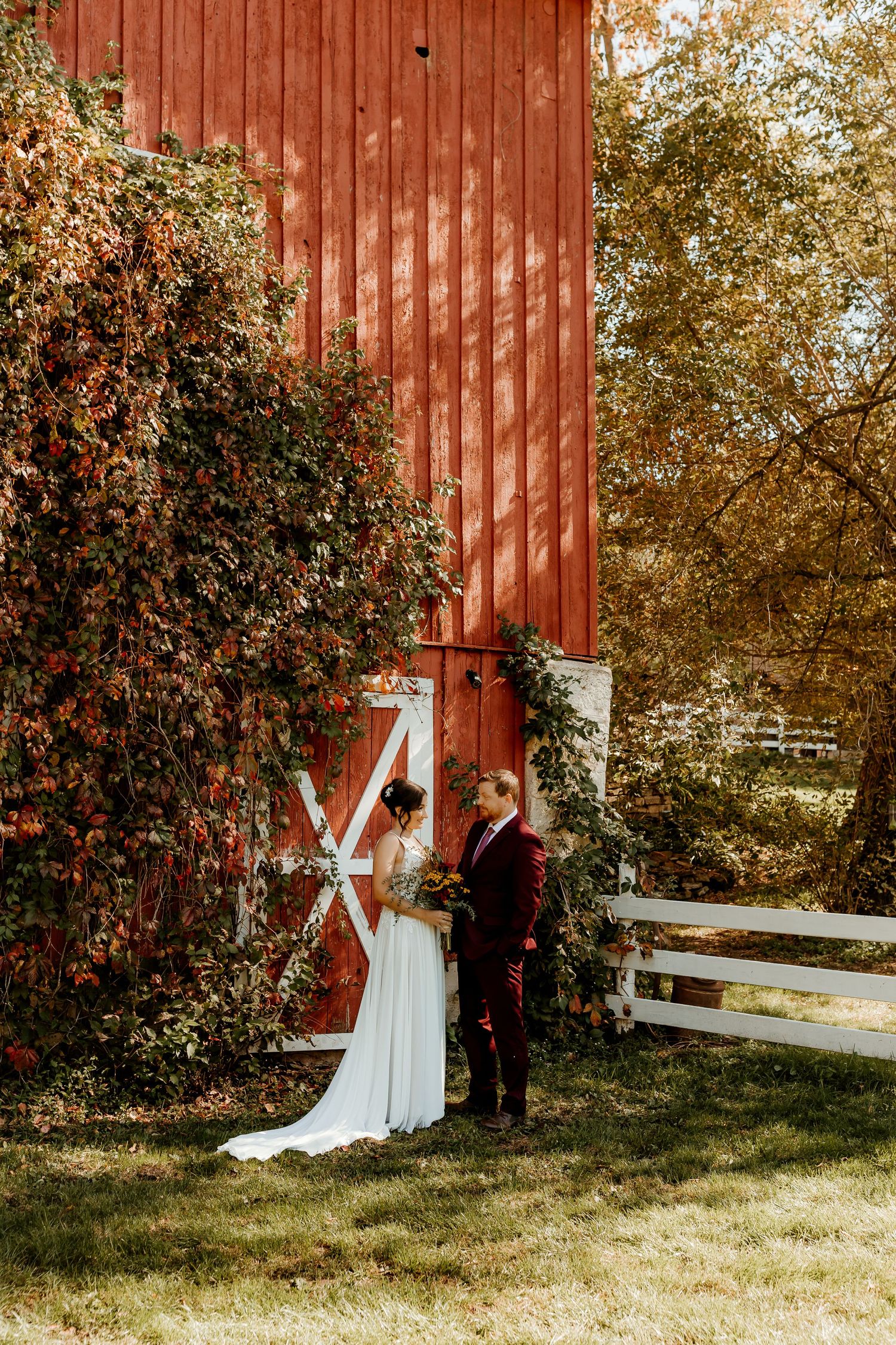 Bride in a white gown and groom in a burgundy suit stand together in front of a red barn with white doors, surrounded by autumn foliage.