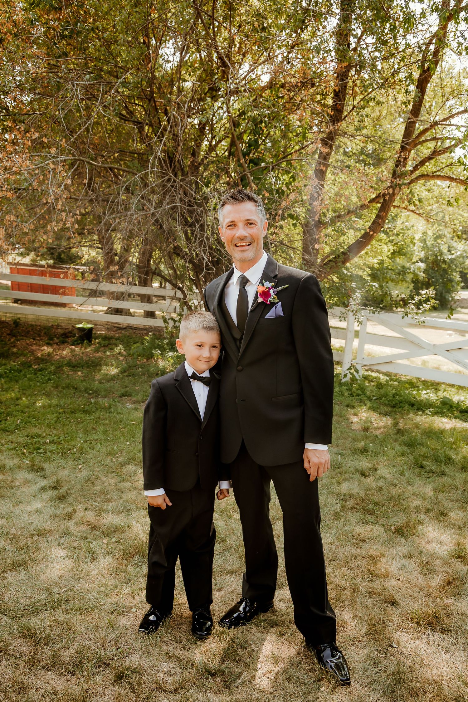 A smiling groom in a black tuxedo with boutonniere stands beside a young boy in matching black tuxedo outdoors.