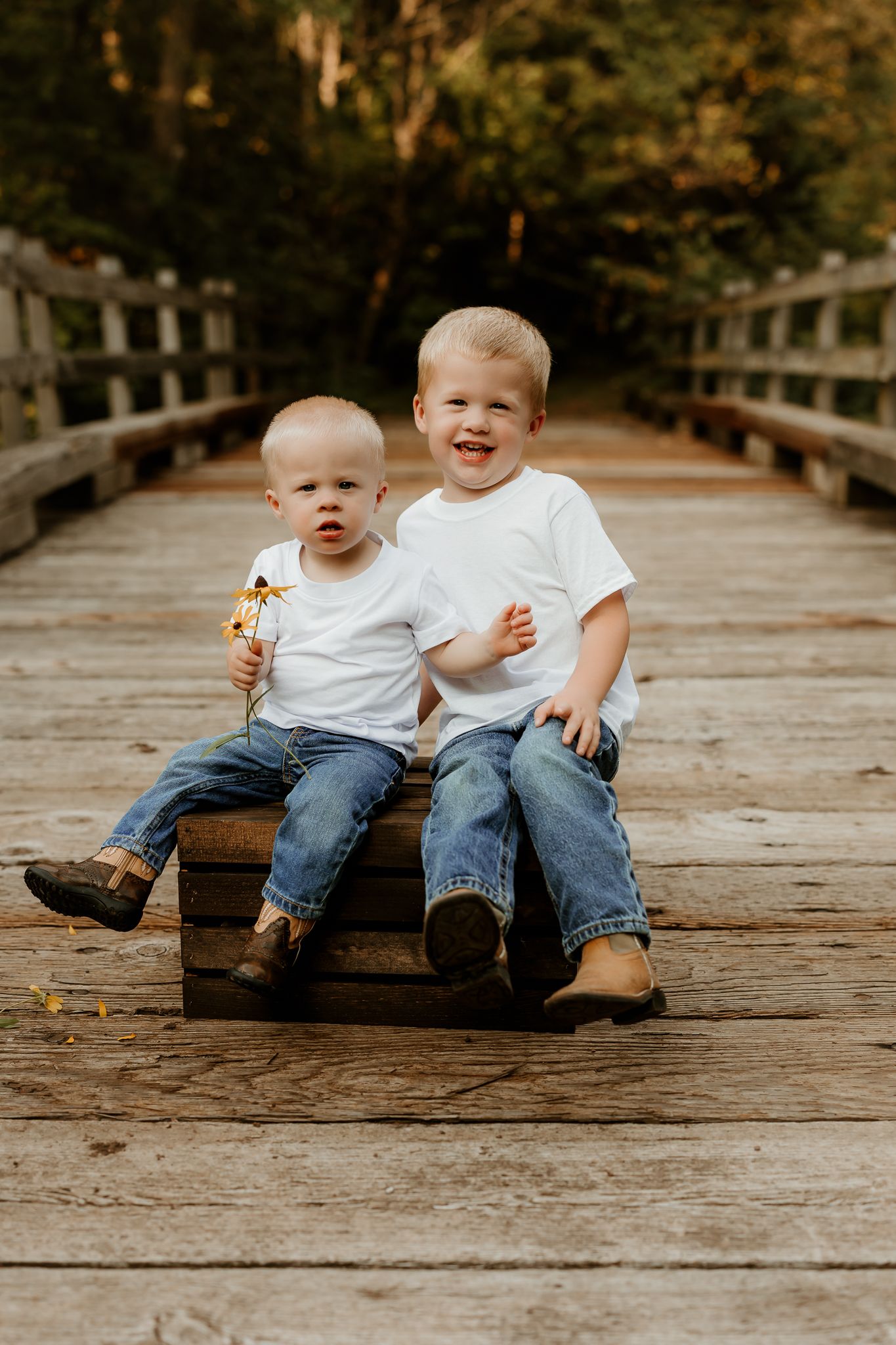 Two young brothers in matching white tees and jeans sit together on a wooden bridge, one holding a flower.