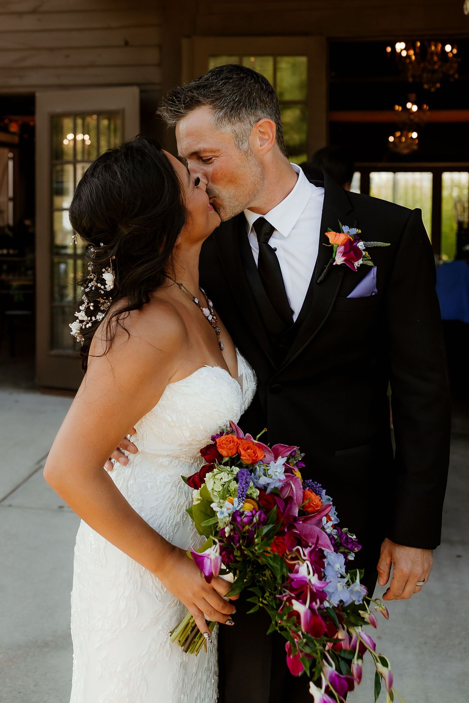 Bride and groom share a romantic kiss, she holds a vibrant bouquet of purple, orange, and red flowers, both dressed in wedding attire.