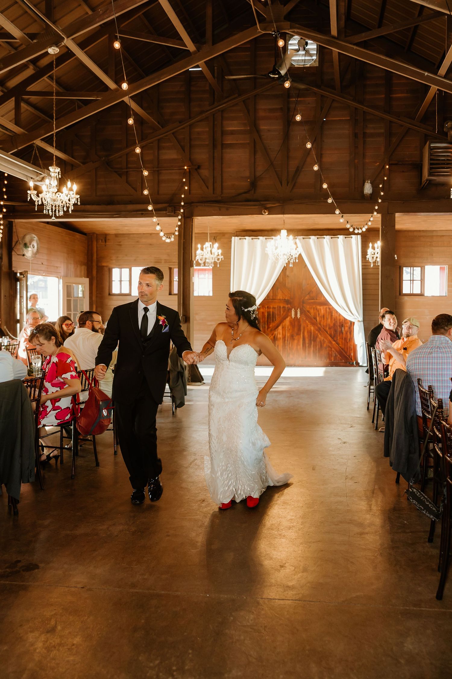 Bride and groom walk hand-in-hand through rustic barn wedding reception with chandeliers and string lights.