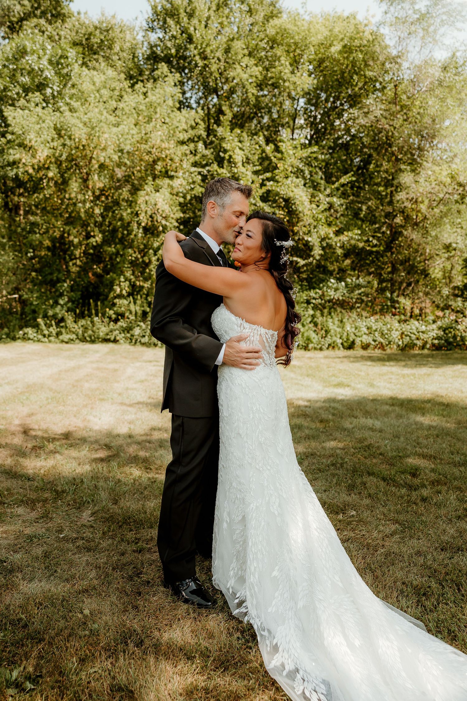 A bride and groom share a romantic embrace outdoors, surrounded by lush green trees on their wedding day.