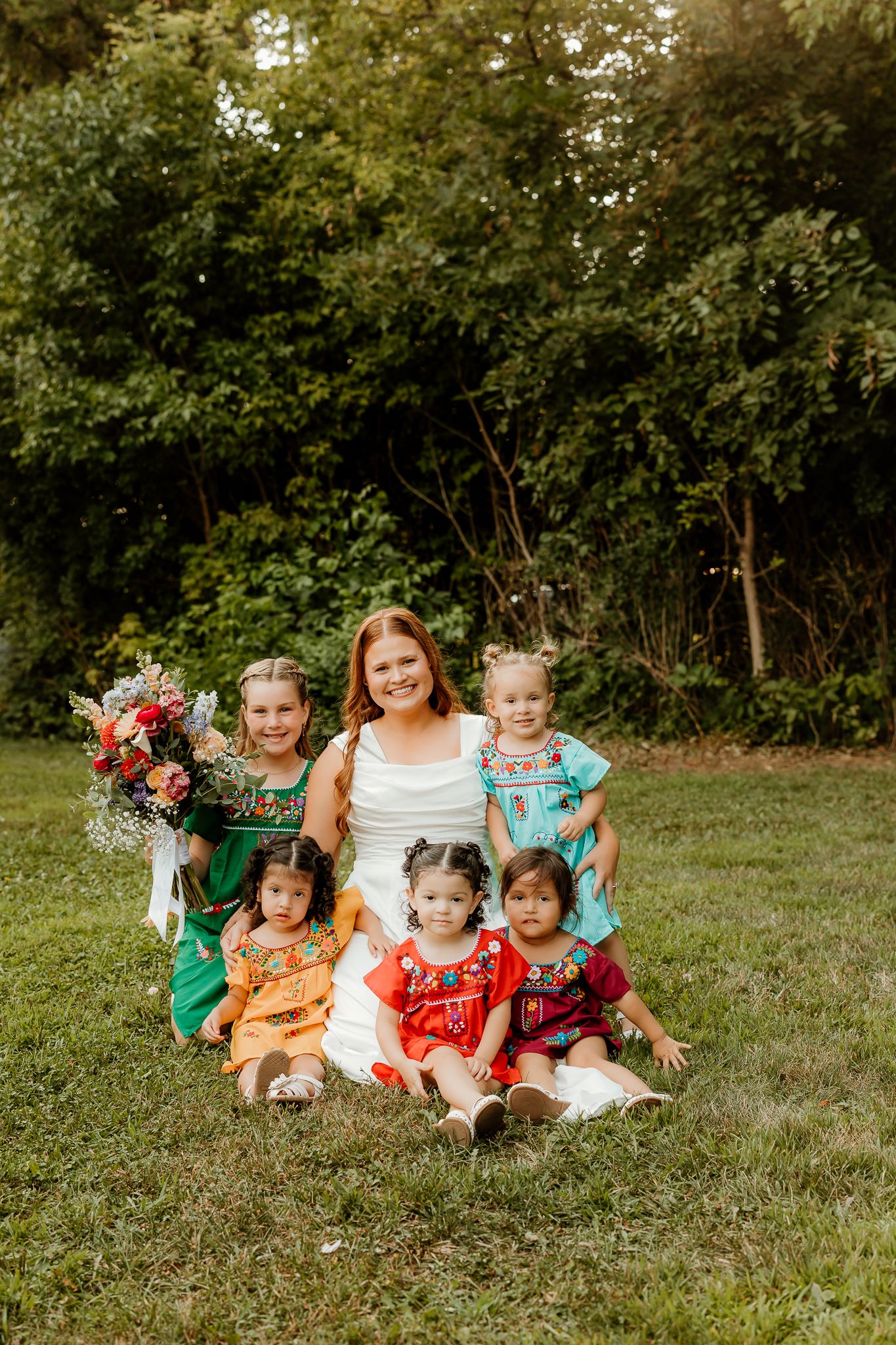 A bride in white poses with five young girls in colorful embroidered dresses on green grass outdoors.