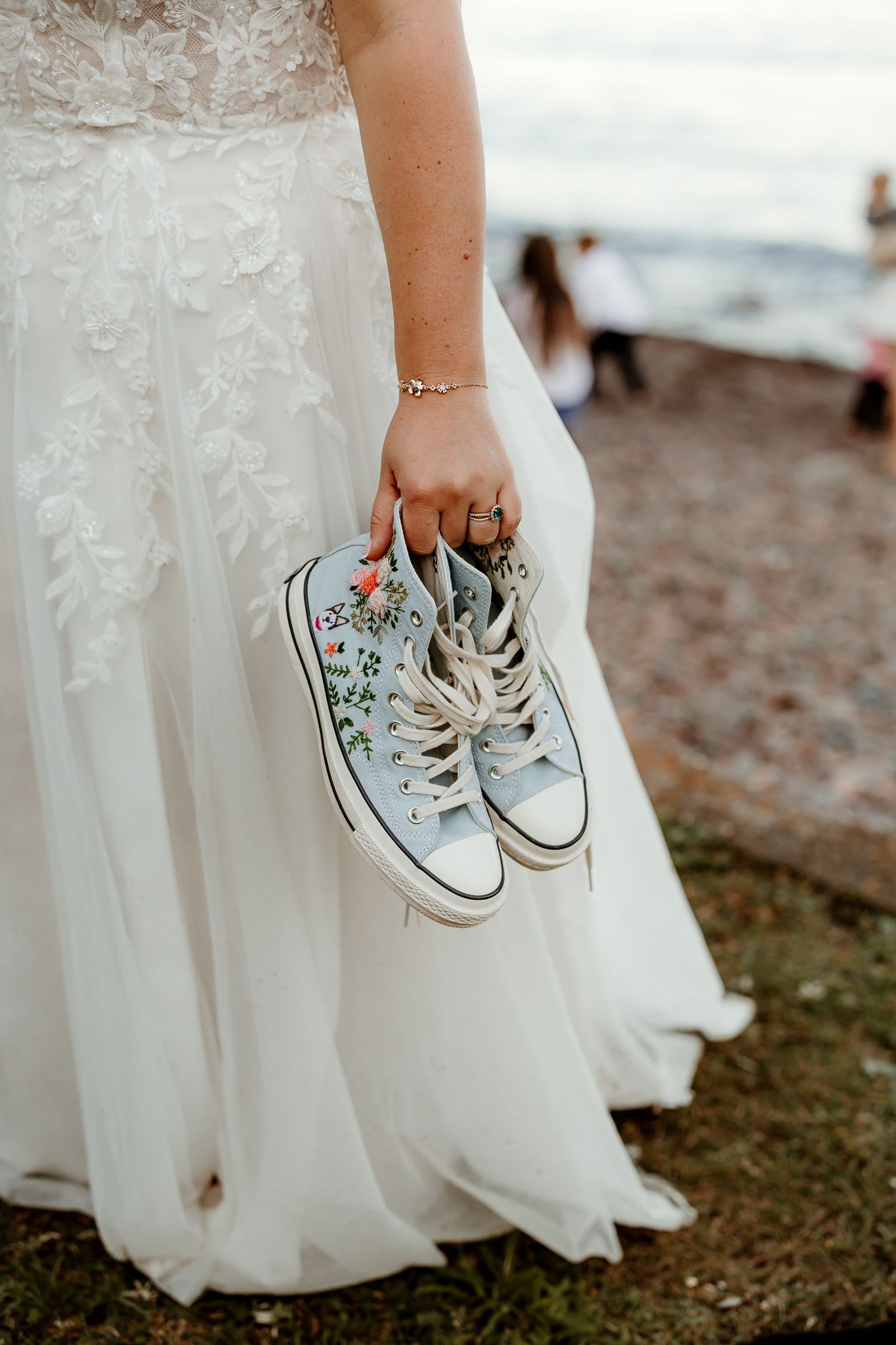 Bride in white lace gown holding light blue floral Converse sneakers at an outdoor wedding venue.