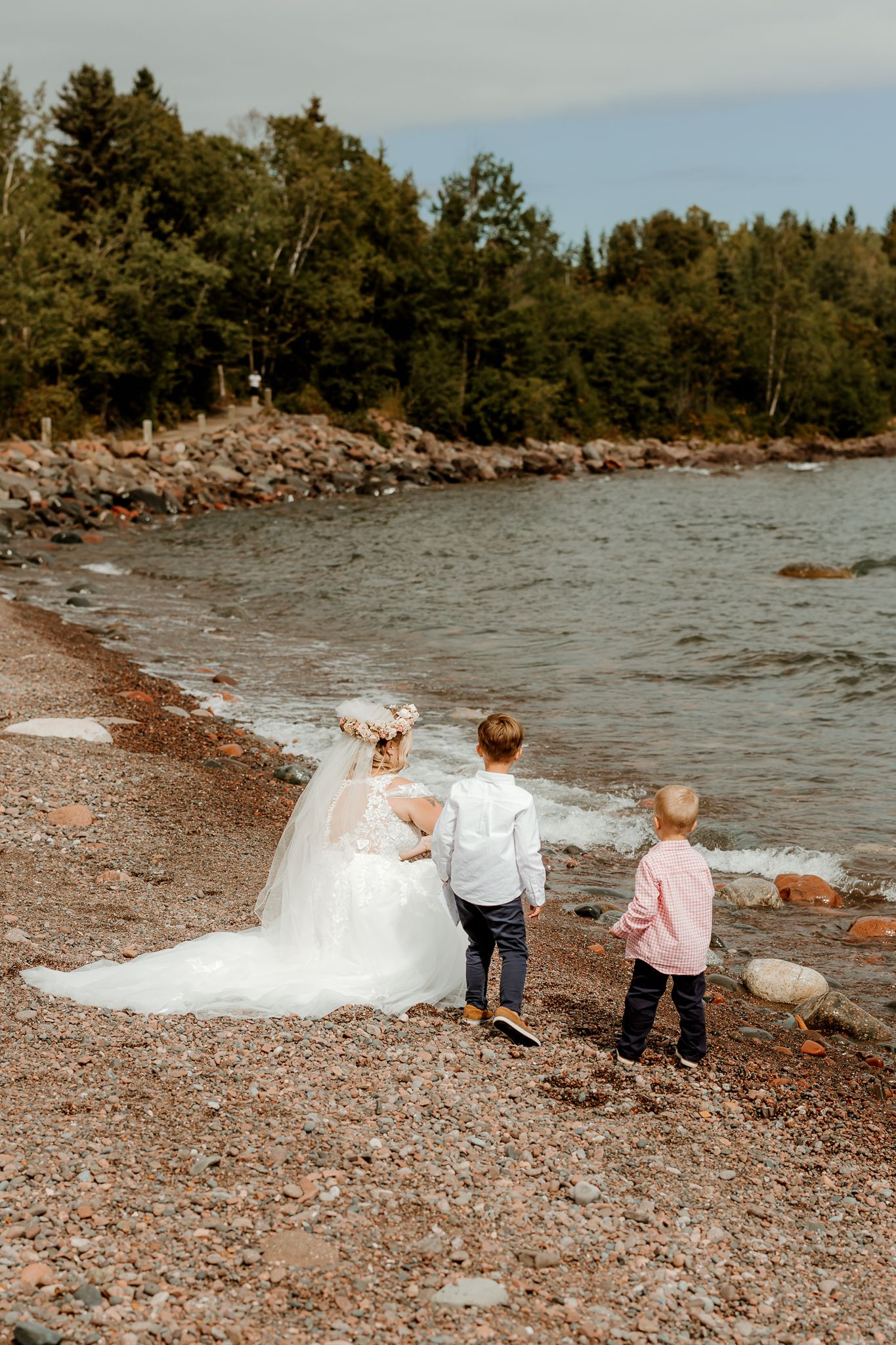 Bride in white wedding gown with veil walks along rocky shoreline with two young boys near forested waterfront.
