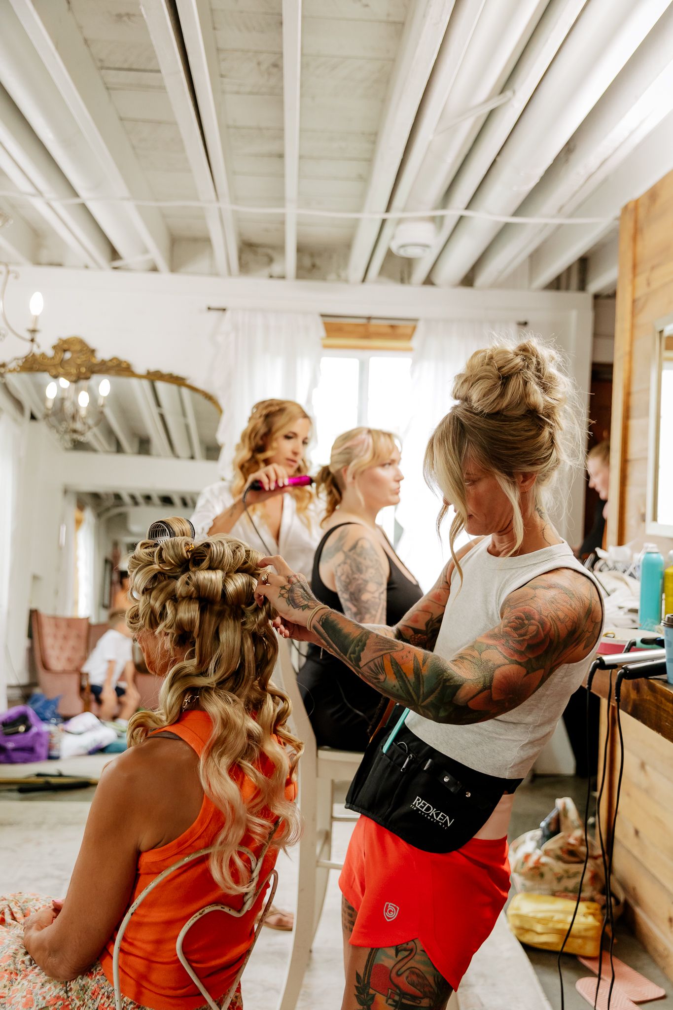 Tattooed hairstylist styling wedding party hair in a bright rustic venue with multiple women getting ready.