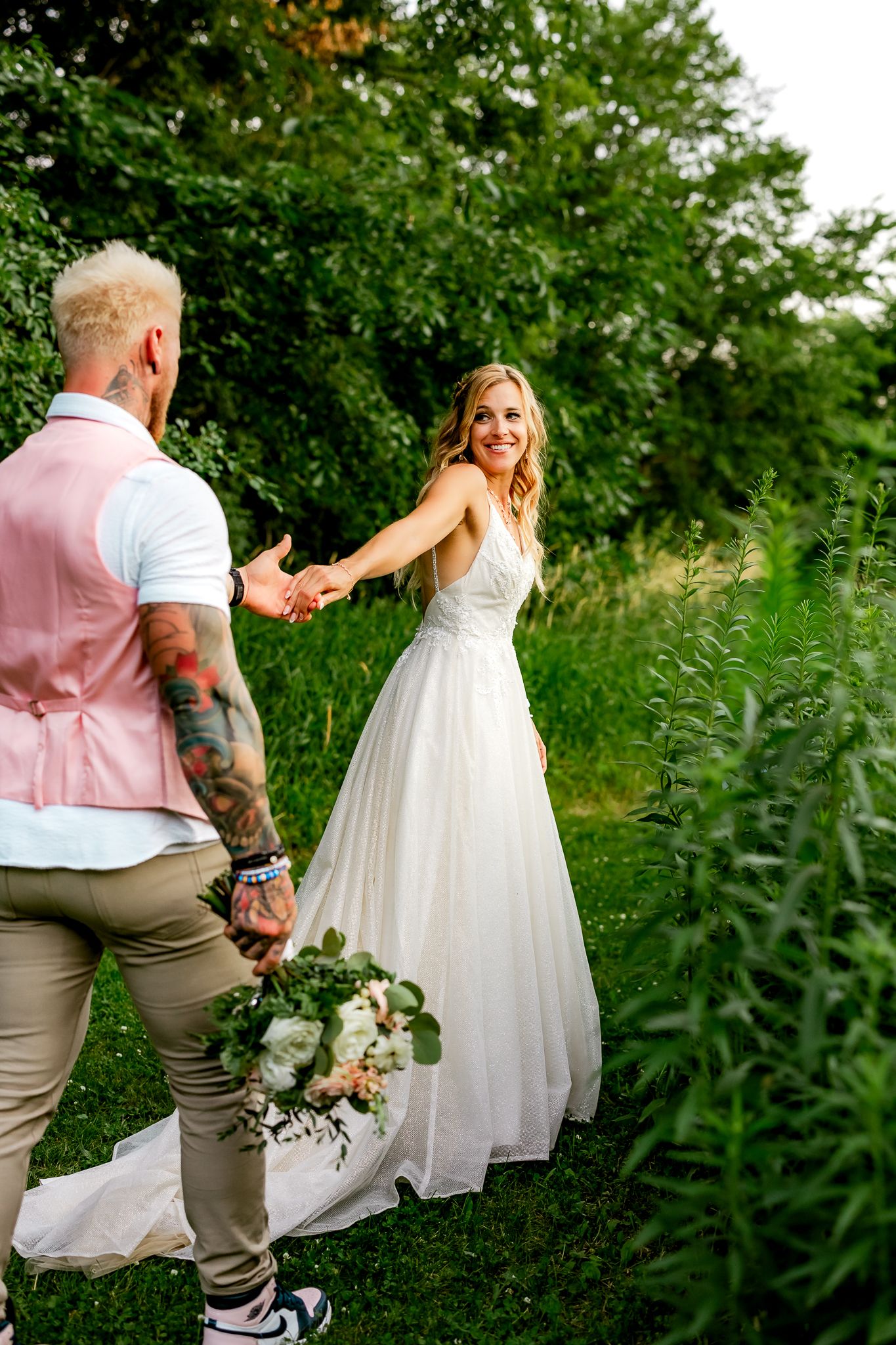 Smiling bride in white gown holds grooms hand in lush green garden during outdoor wedding photo session.