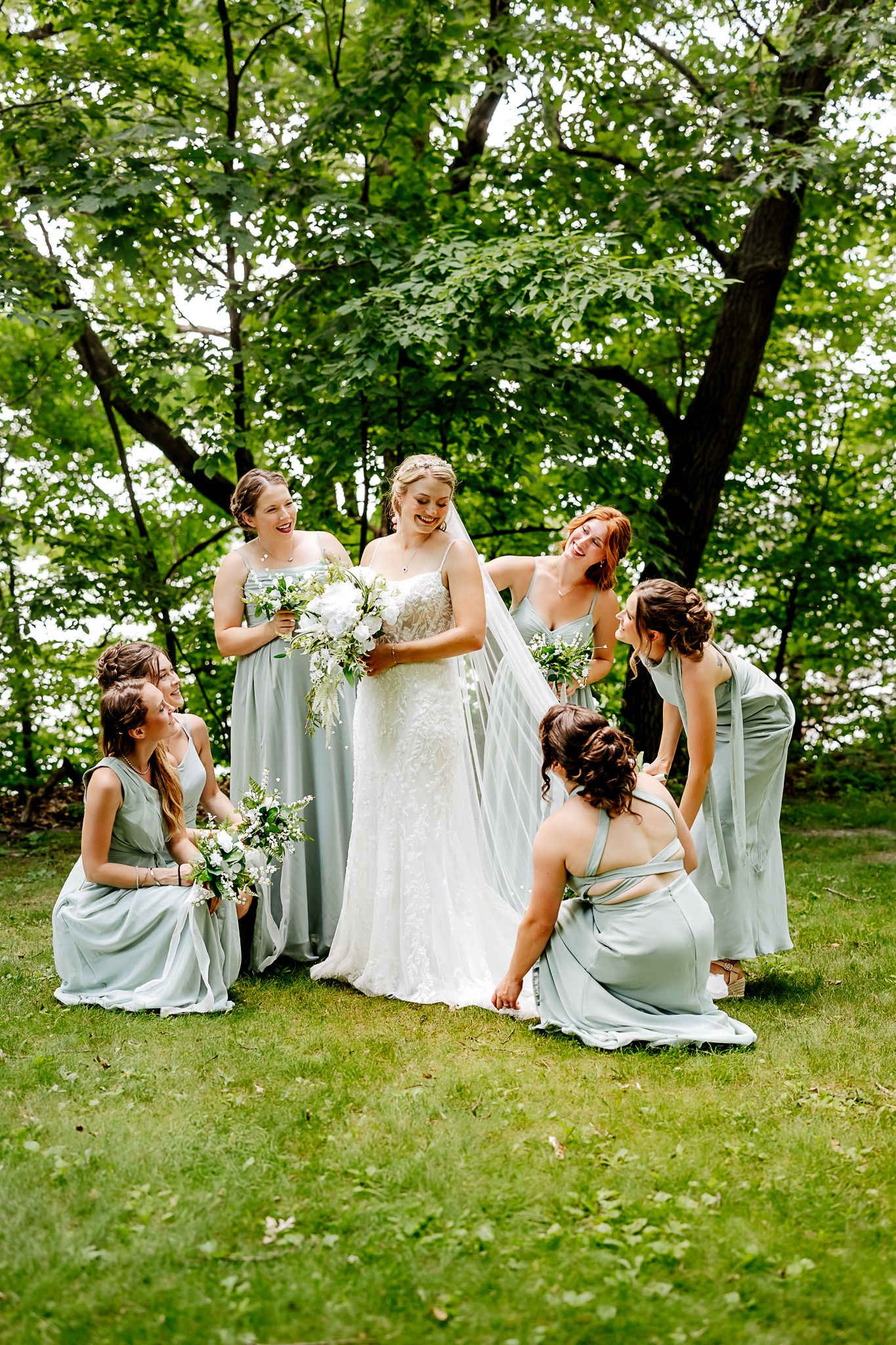 Bride in white gown with bridesmaids in sage green dresses posing outdoors in lush green garden setting.