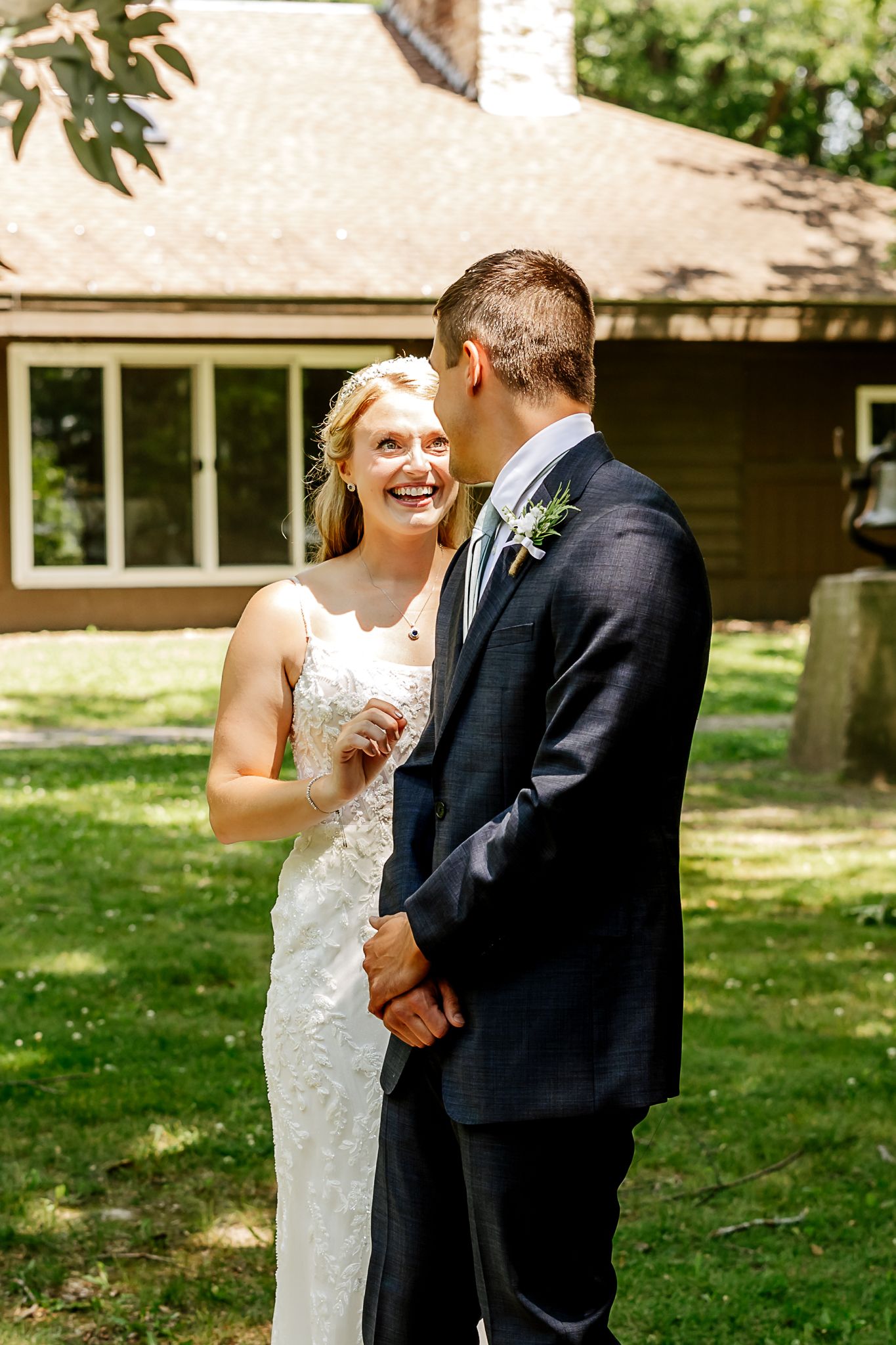A smiling bride in a white lace gown stands with a groom in a navy suit outdoors near a rustic building.