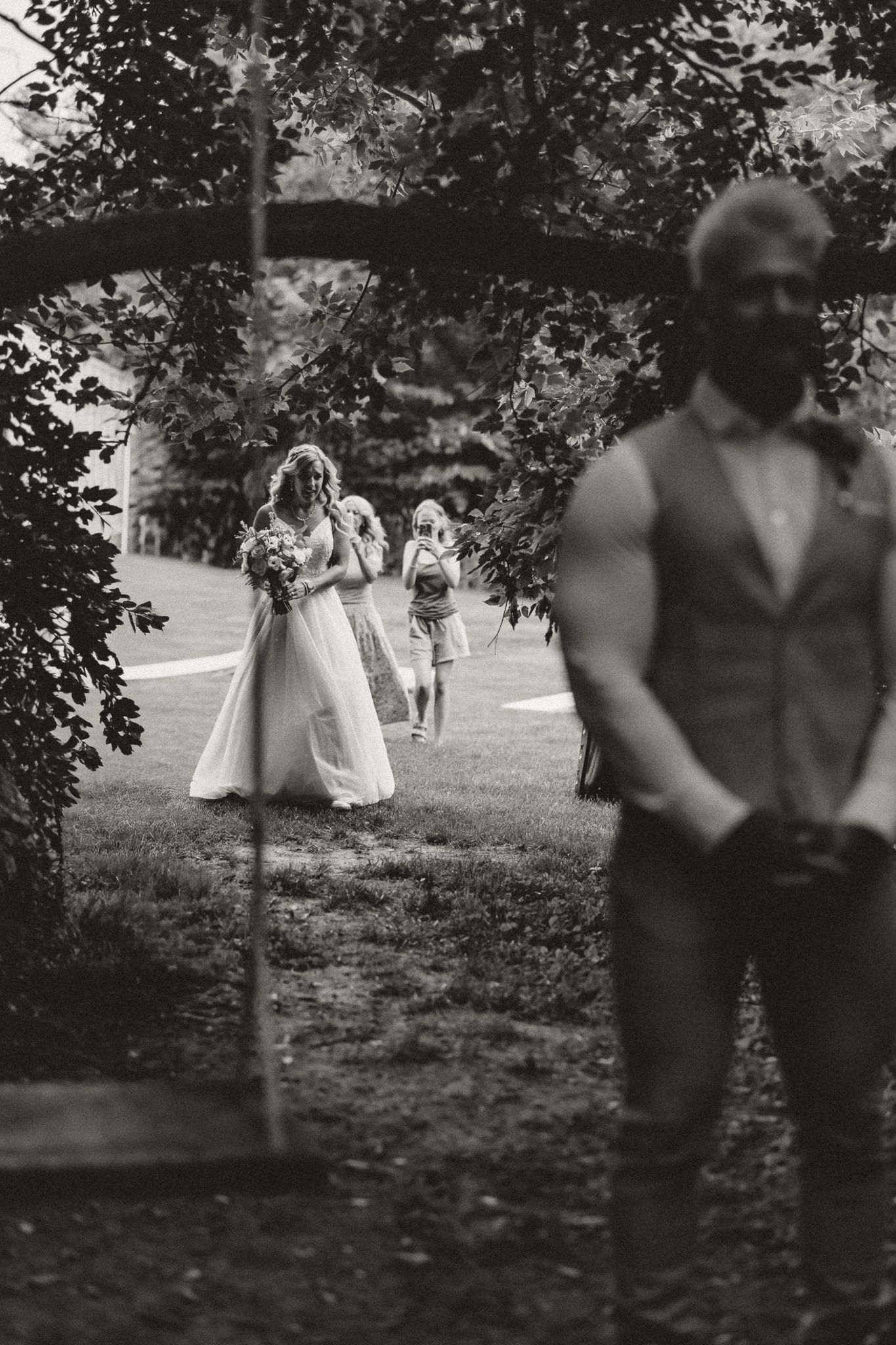 Black and white wedding photo of bride walking through garden arch toward groom in vest, with flower girls following behind.