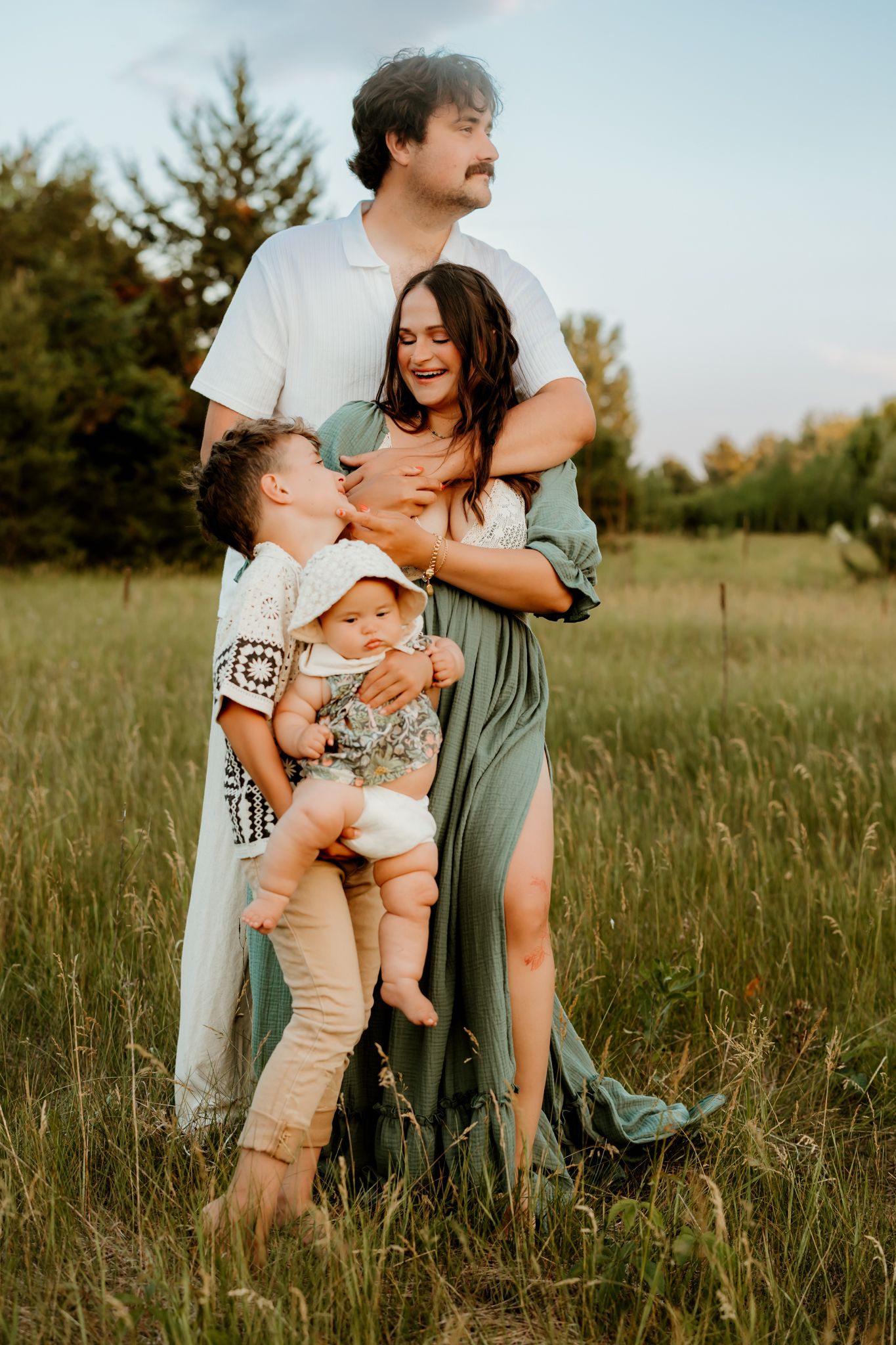 Happy family of four posing in a green meadow, parents embracing while holding their two young children outdoors.