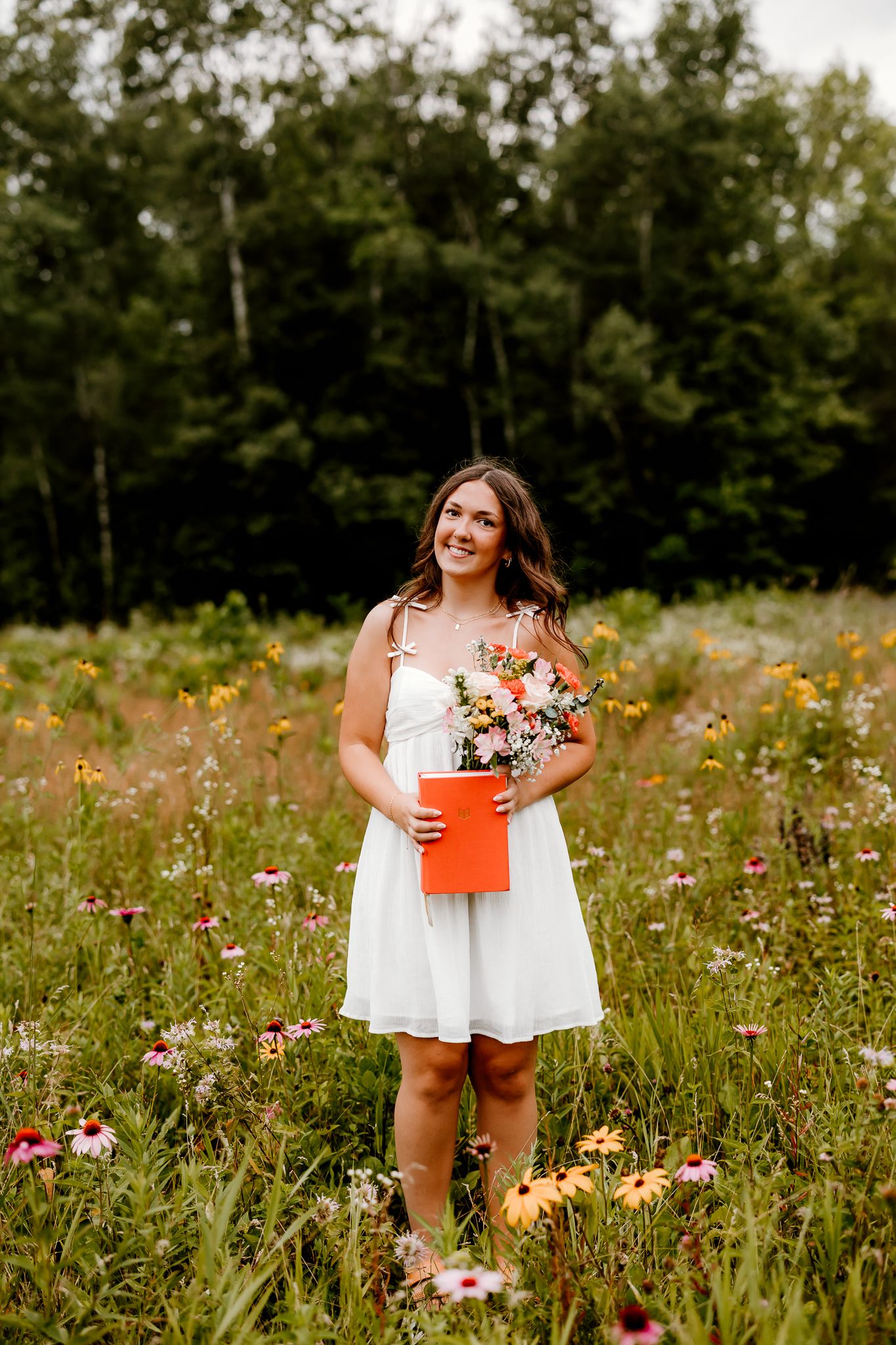 Young woman in white dress holding flowers and orange folder standing in colorful wildflower meadow with trees behind.