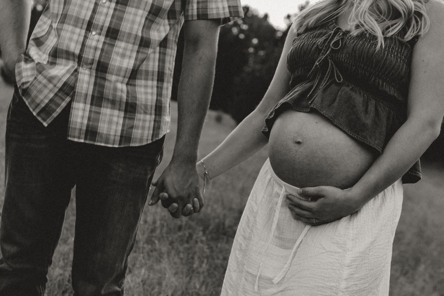 Black and white maternity photo of couple holding hands in field, pregnant woman cradling baby bump outdoors.