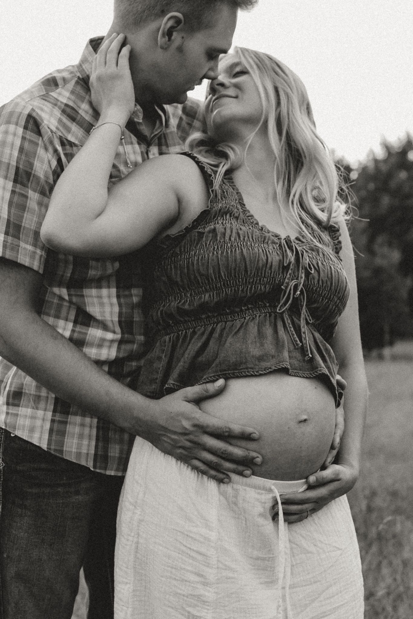 Black and white maternity photo of a couple embracing outdoors, man kissing woman's forehead while holding her baby bump.