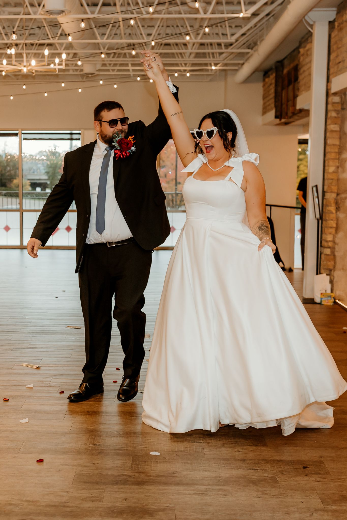 Joyful bride in a white gown and heart sunglasses raises hands with her groom in a dark suit and red boutonniere at their wedding.