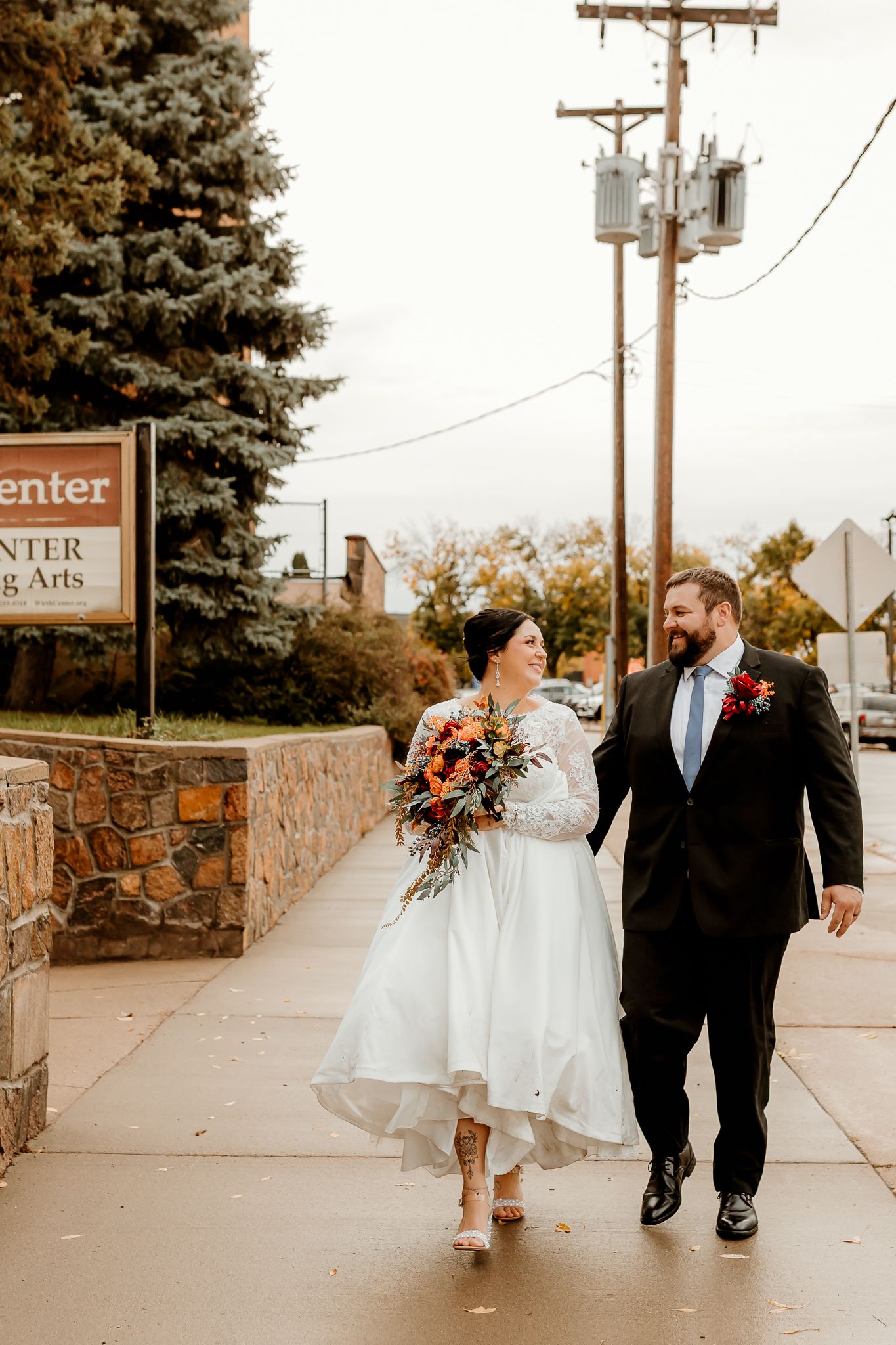 Bride in a tea-length white gown and groom in a dark suit hold hands and walk down a sidewalk in autumn, she carrying a colorful bouquet.