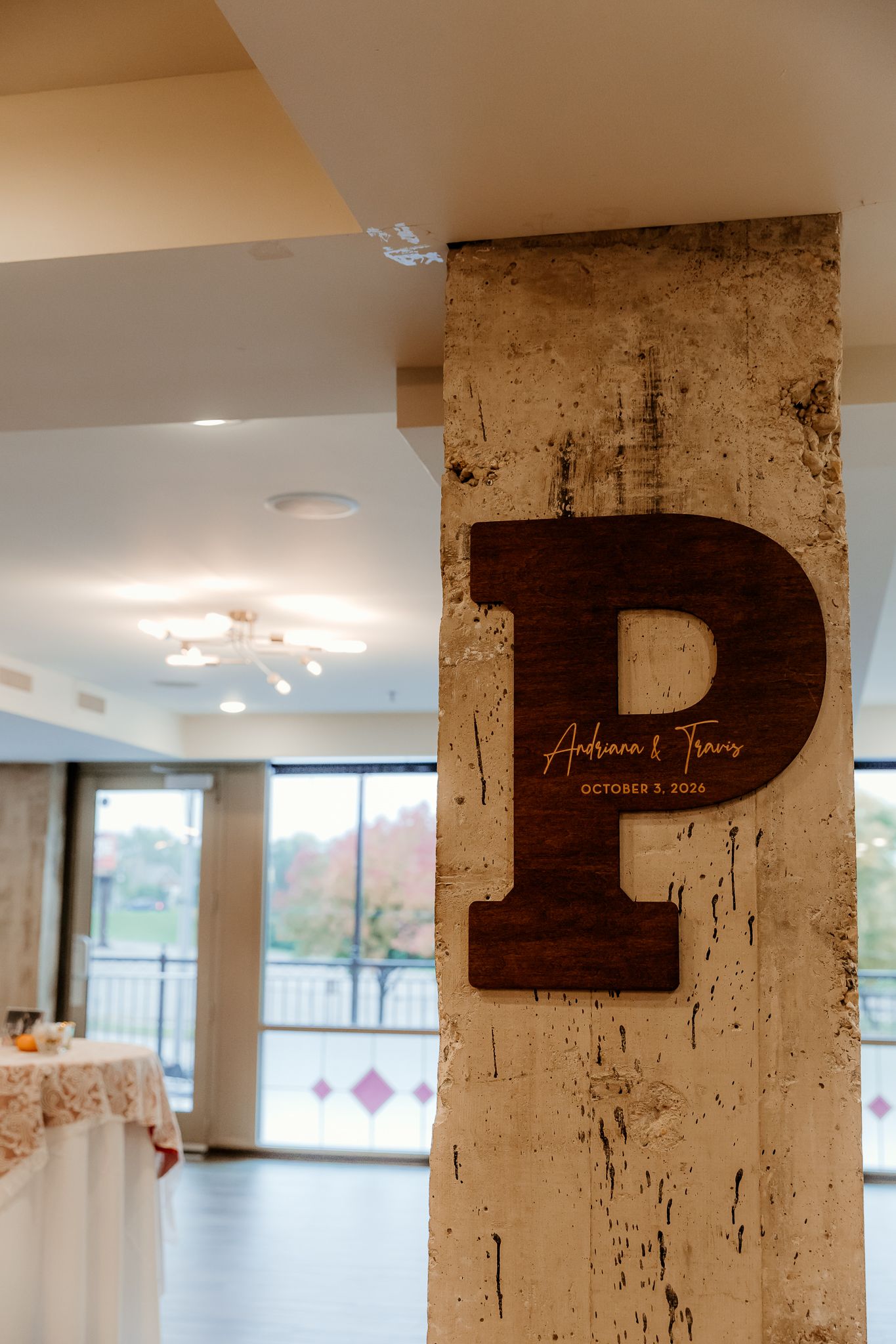 Large dark wooden letter P with the names Andriana and Travis and date October 2026 mounted on a stone pillar at a bright wedding venue.