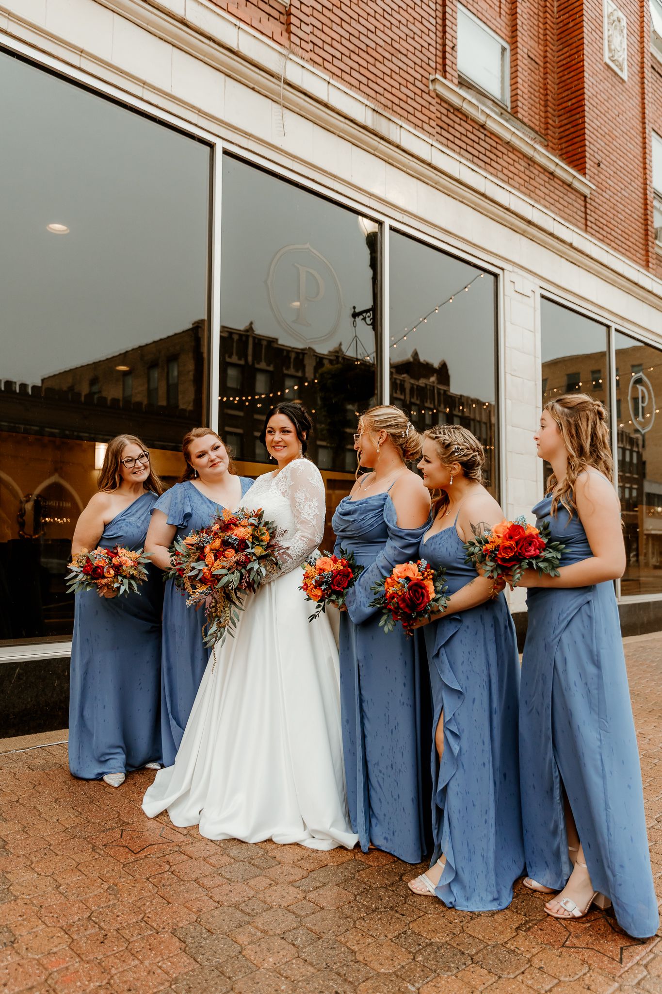 Bride in a white lace gown stands among six bridesmaids in dusty blue dresses holding rust and orange bouquets outside a brick building.