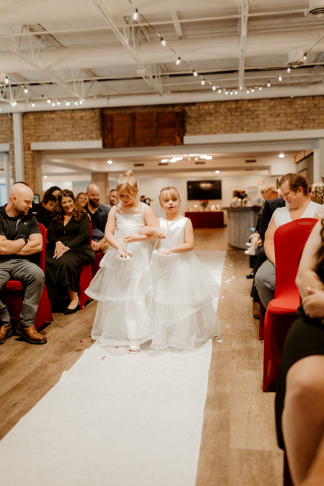 Two flower girls in white tulle dresses walk down a white aisle runner at an indoor wedding ceremony as seated guests look on.