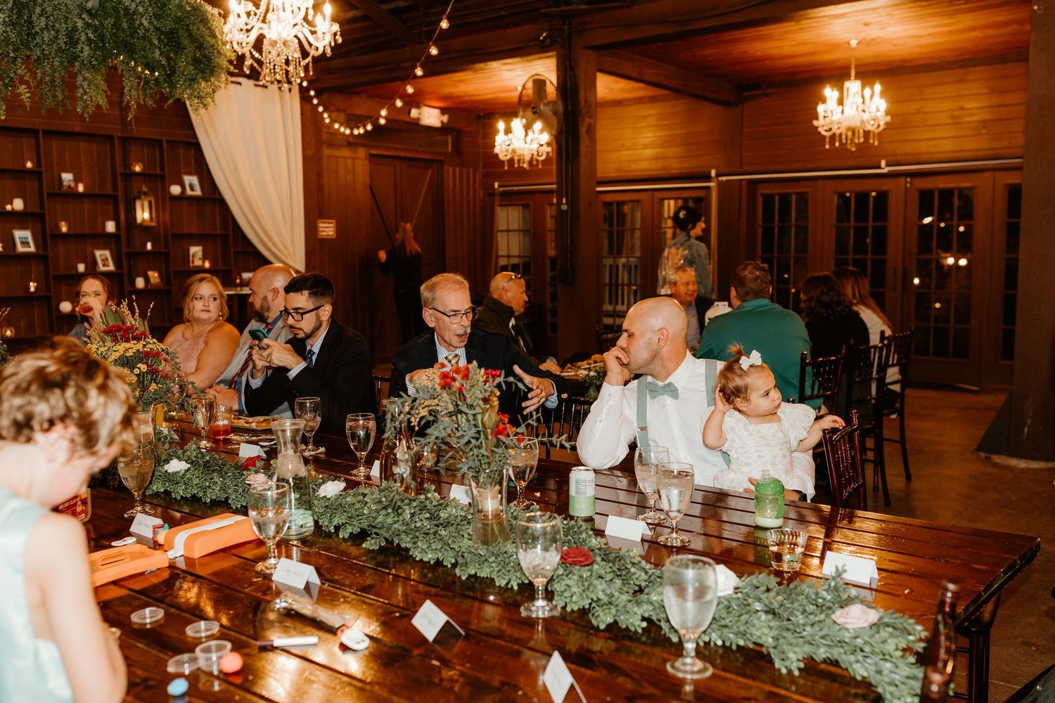 Wedding guests seated at a long wooden banquet table with greenery garland and candles in a rustic venue lit by chandeliers.