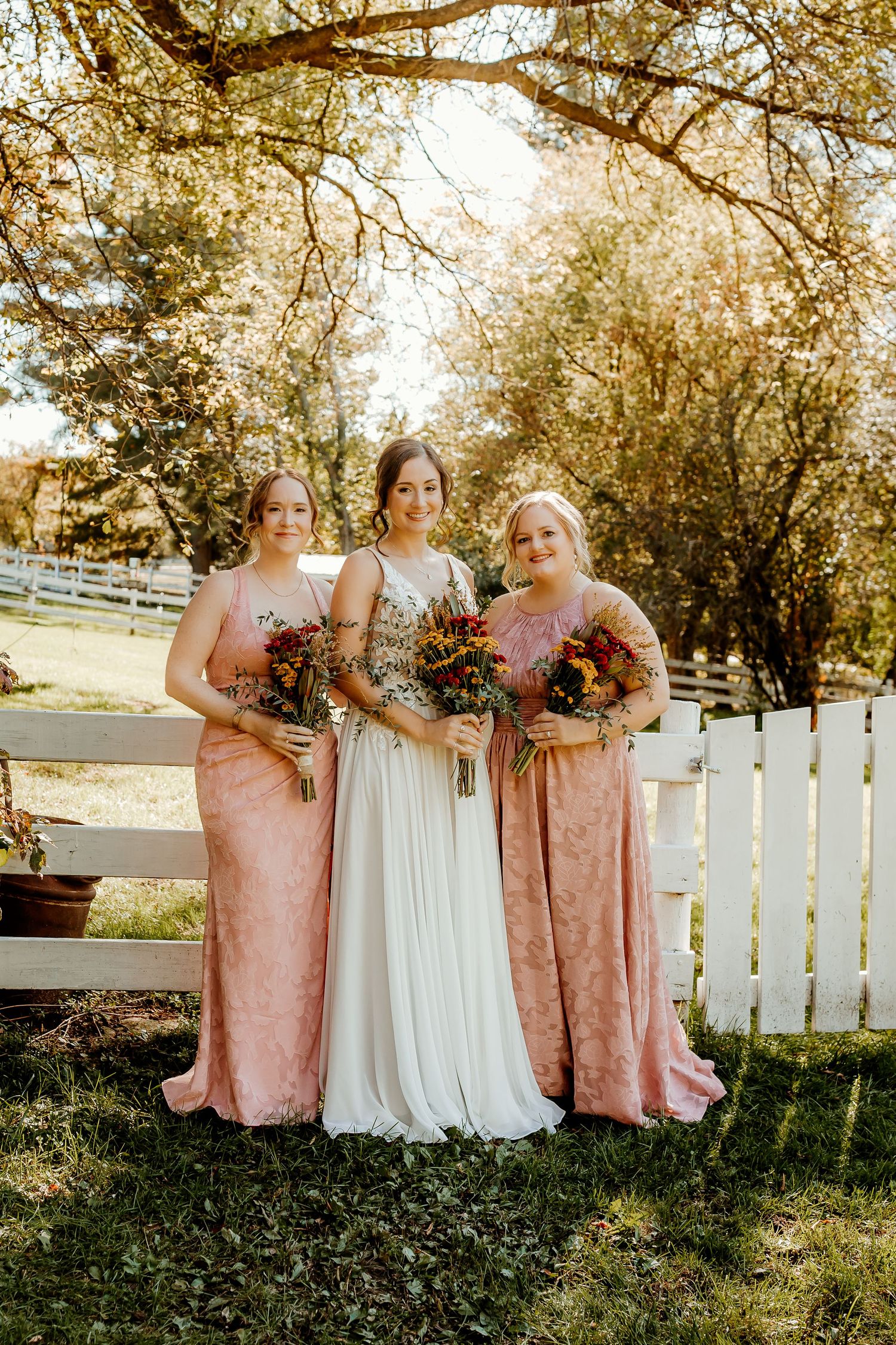 Bride in a white boho gown stands between two bridesmaids in blush pink dresses, all holding colorful bouquets by a white fence.