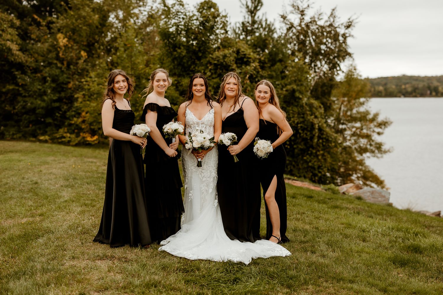 Bride in a white wedding gown stands with four bridesmaids in black dresses holding white bouquets on a grassy lawn by a lake.