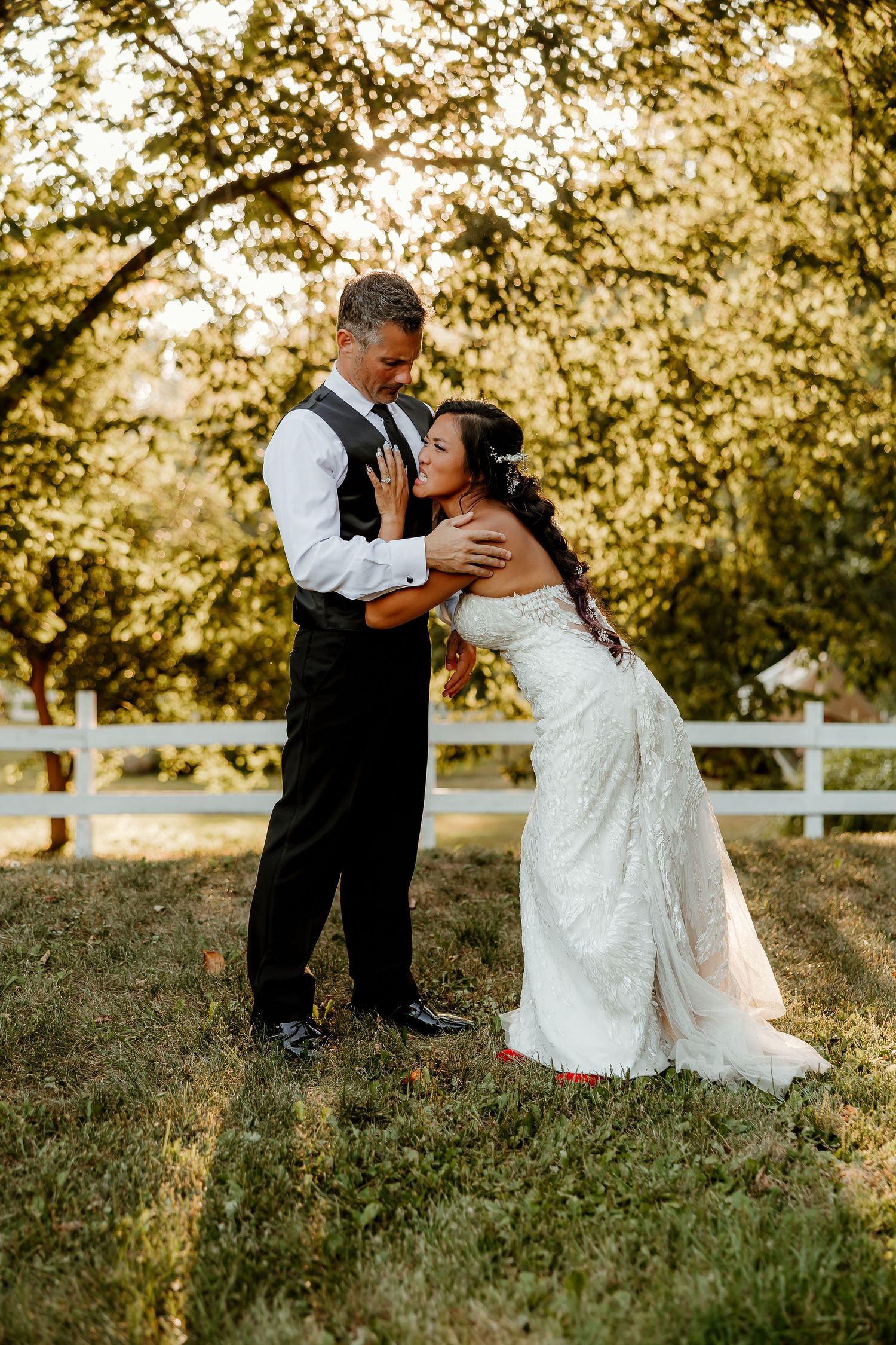 Bride and groom share a romantic moment outdoors near a white fence, surrounded by golden sunlit trees.