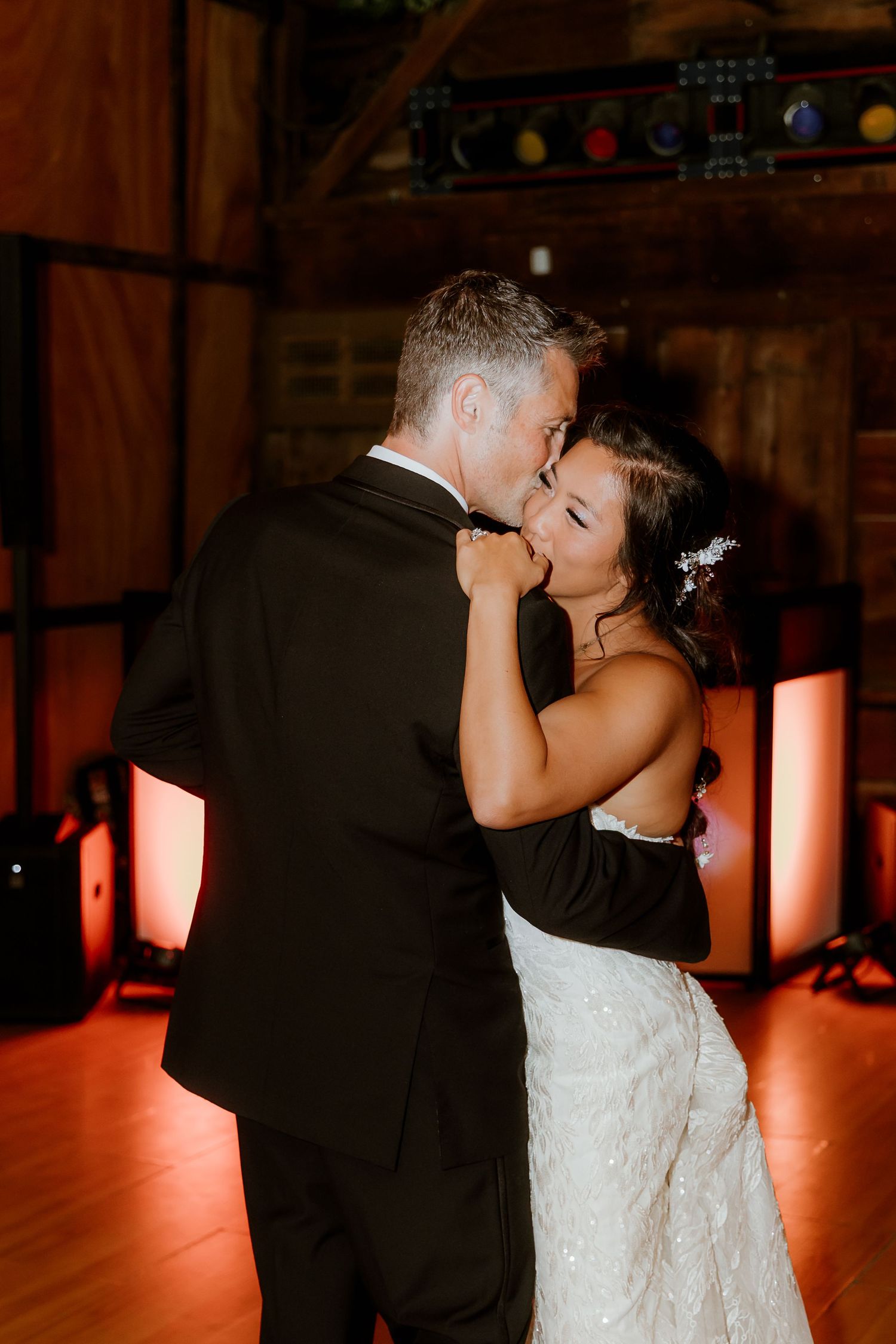 Bride and groom share their first dance, embracing closely in a warmly lit barn wedding reception venue.