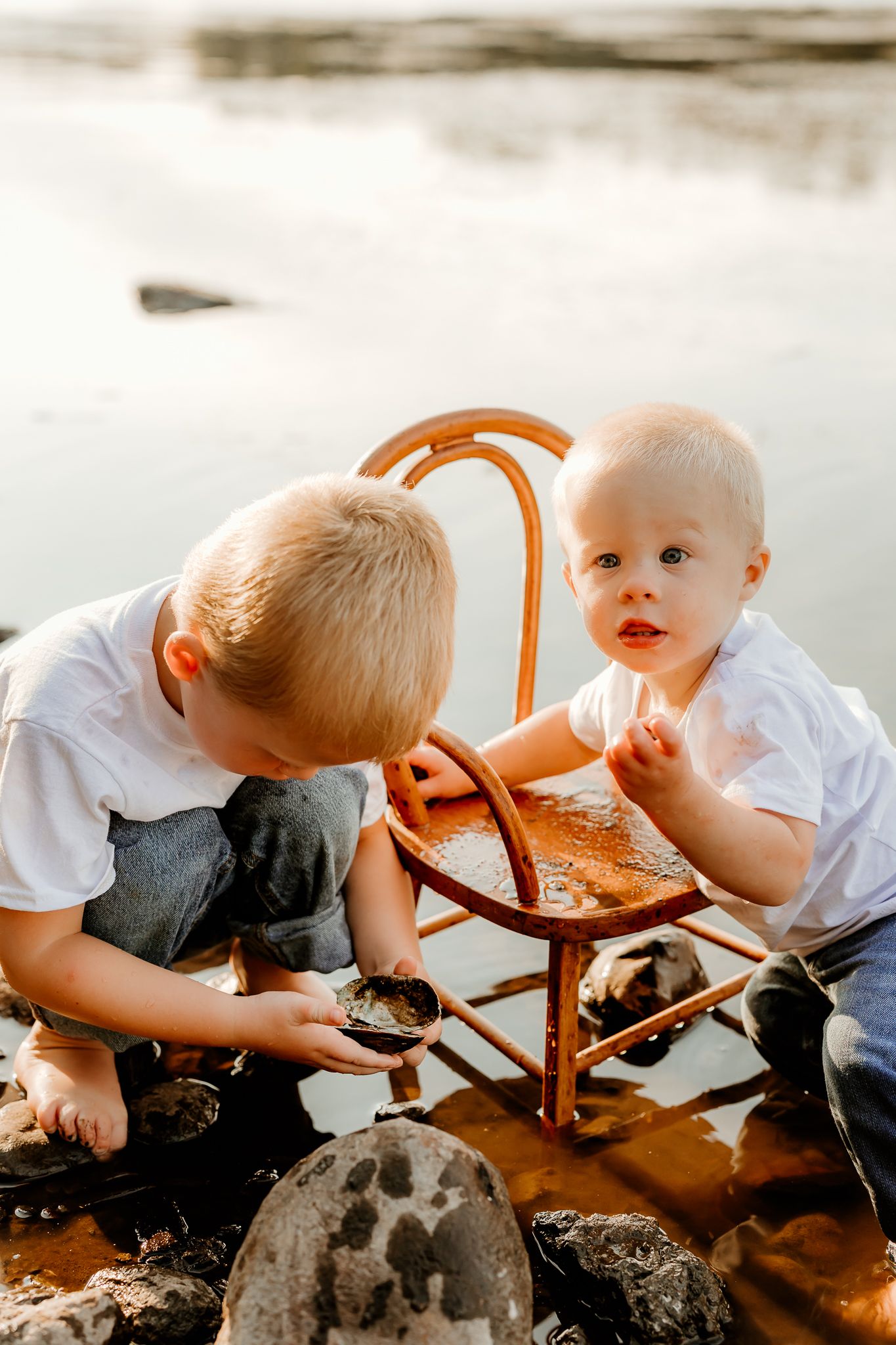 Two young boys in white shirts playing with a wooden chair in shallow water near rocky shoreline.
