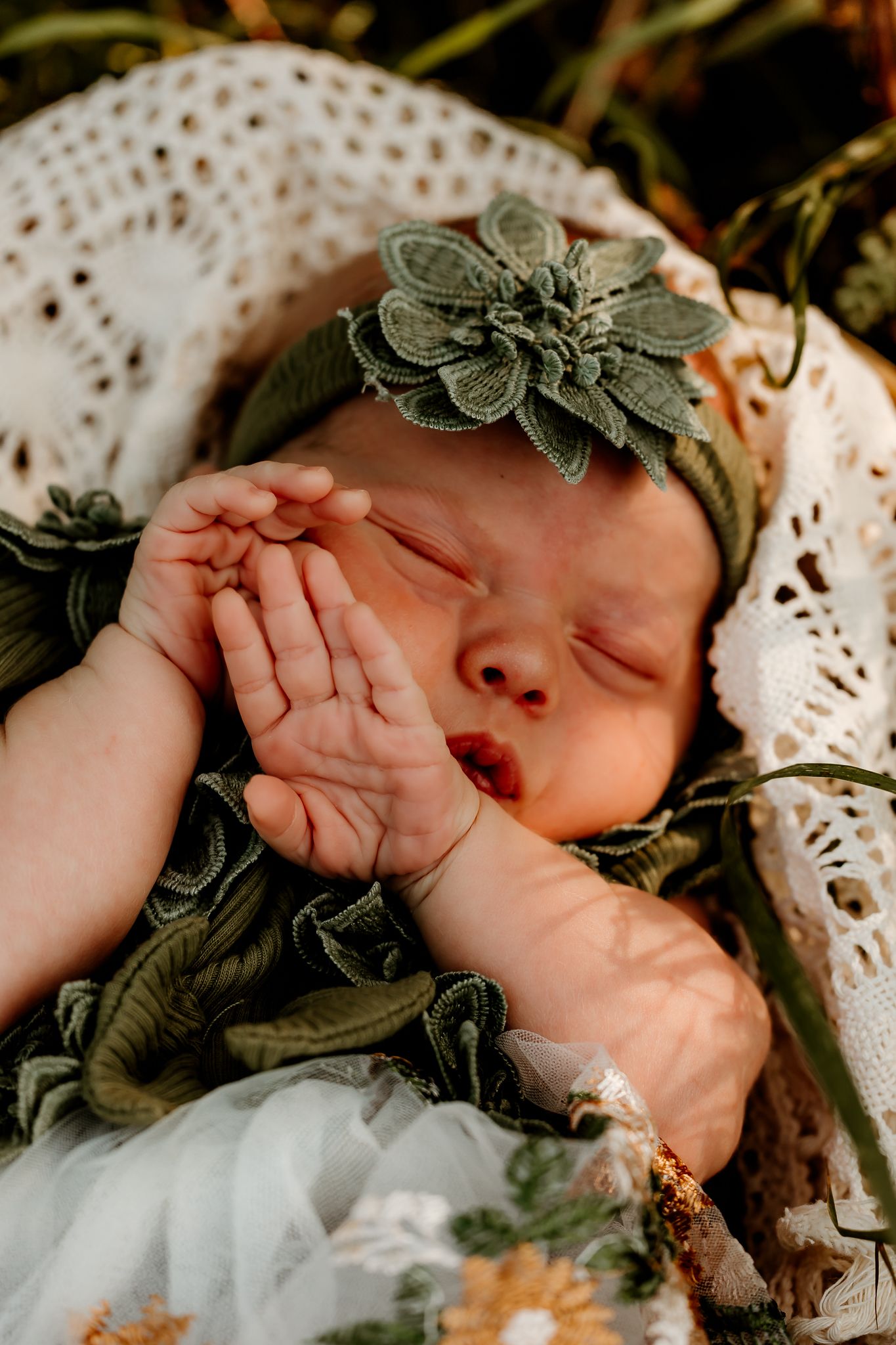 Sleeping newborn baby in green floral outfit and matching headband, resting peacefully in a white lace basket outdoors.