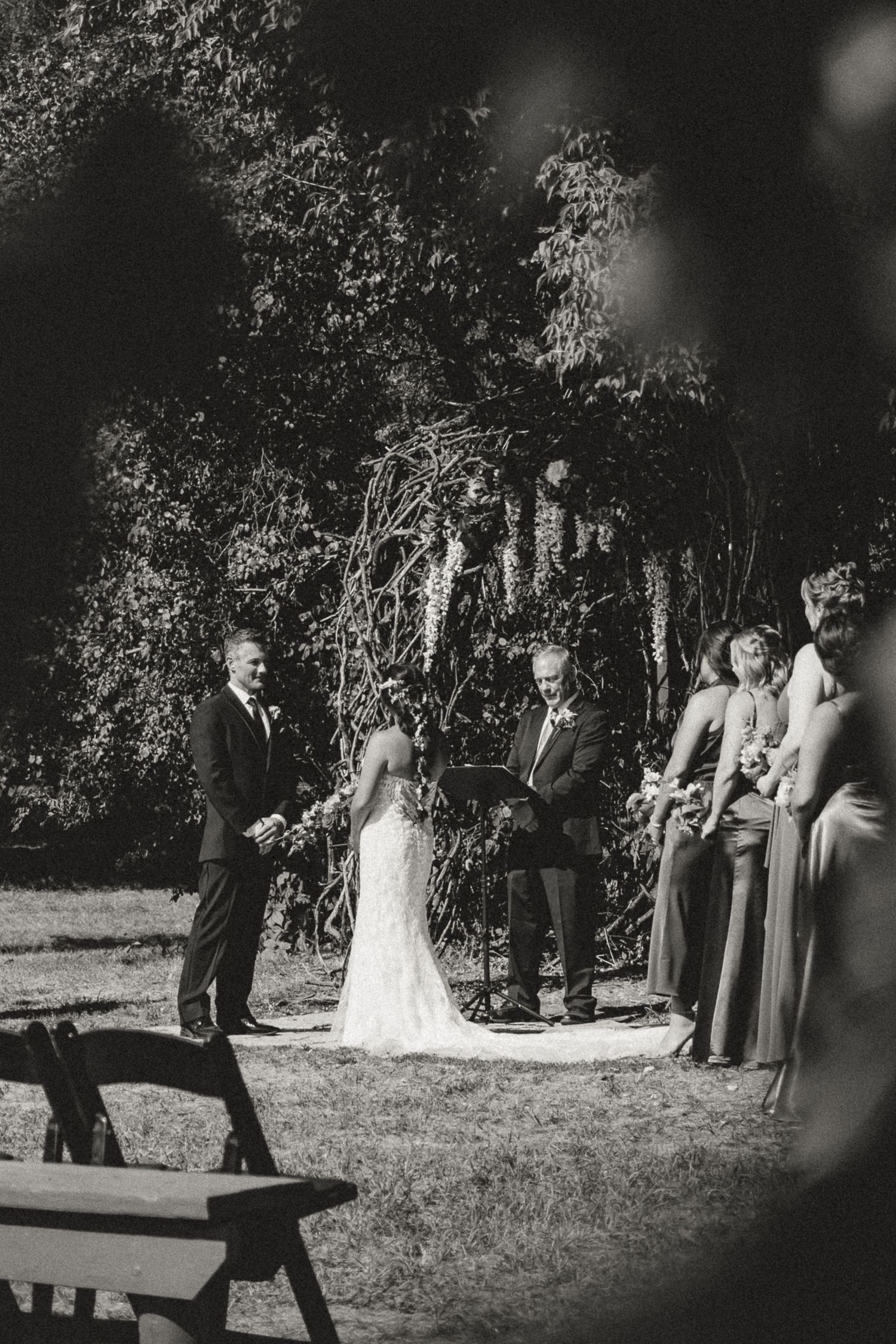 Black and white outdoor wedding ceremony with bride, groom, and officiant standing beneath a floral arch surrounded by lush trees.
