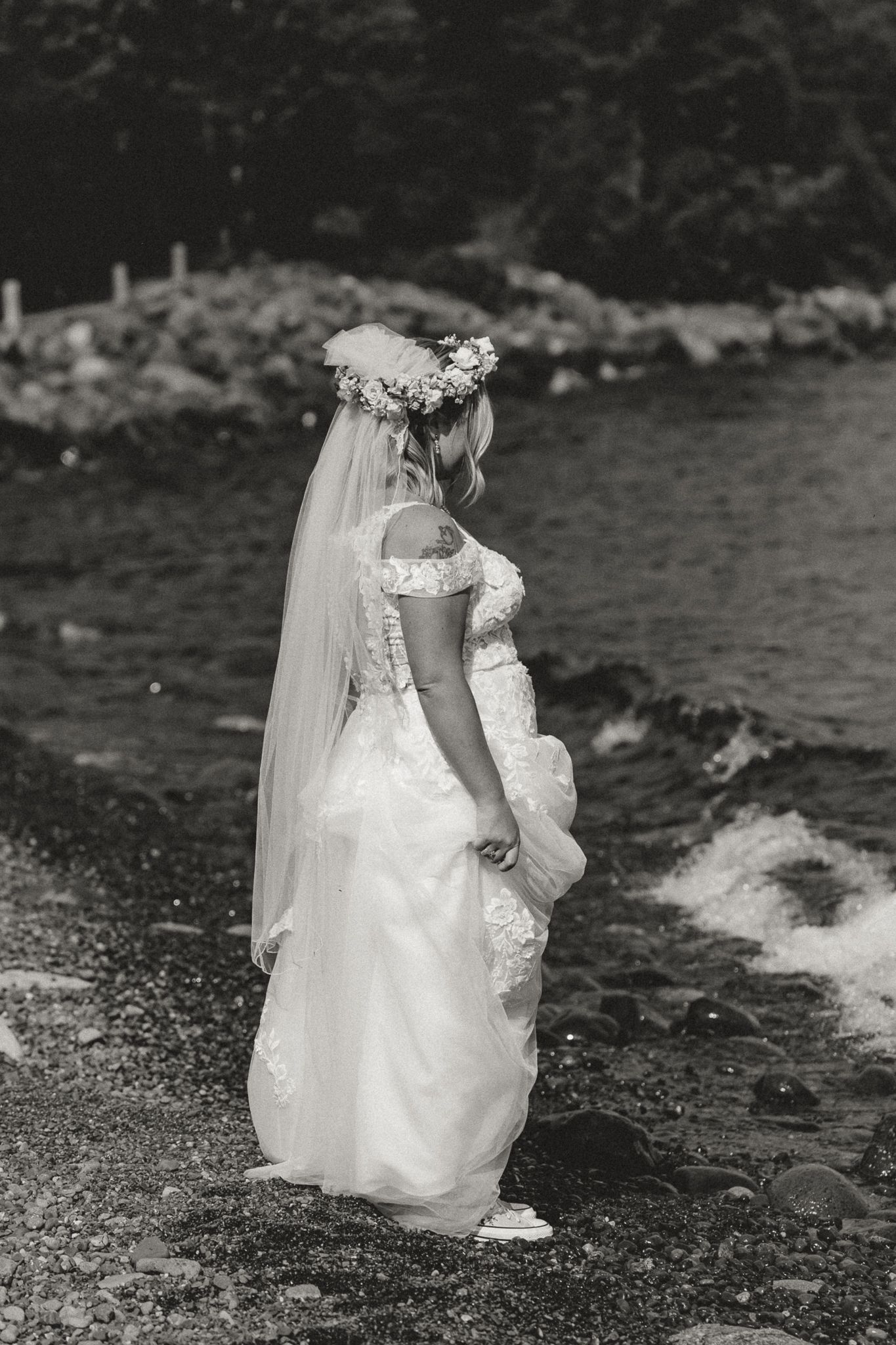 Bride in white lace gown and floral crown stands by rocky shoreline, holding dress, black and white photo.