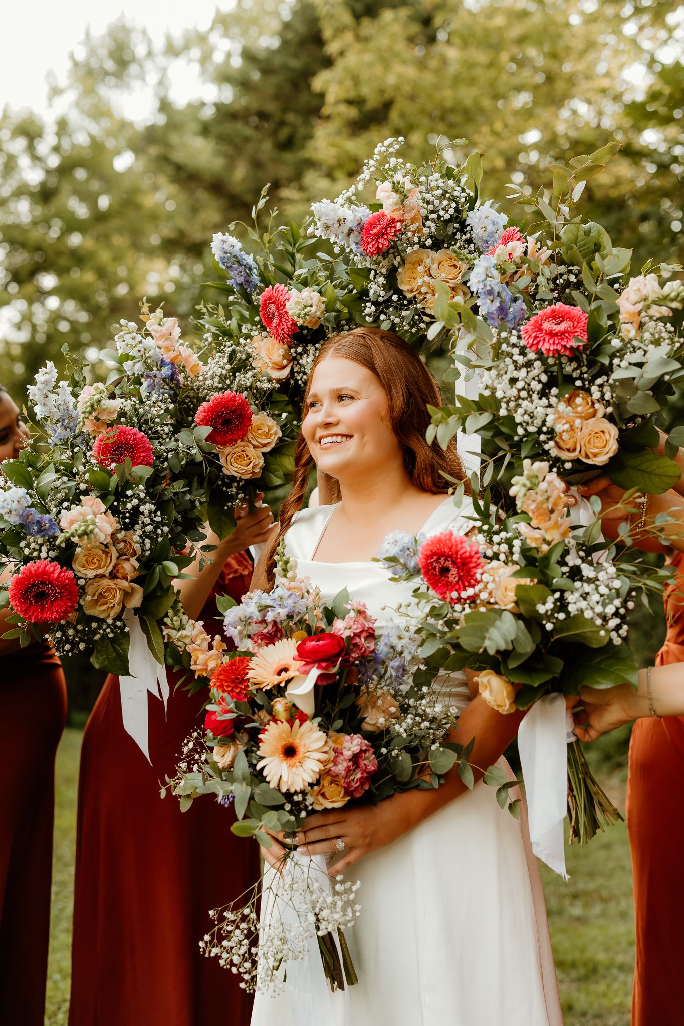 Smiling bride in white dress surrounded by large colorful floral bouquets with bridesmaids in burgundy dresses outdoors.