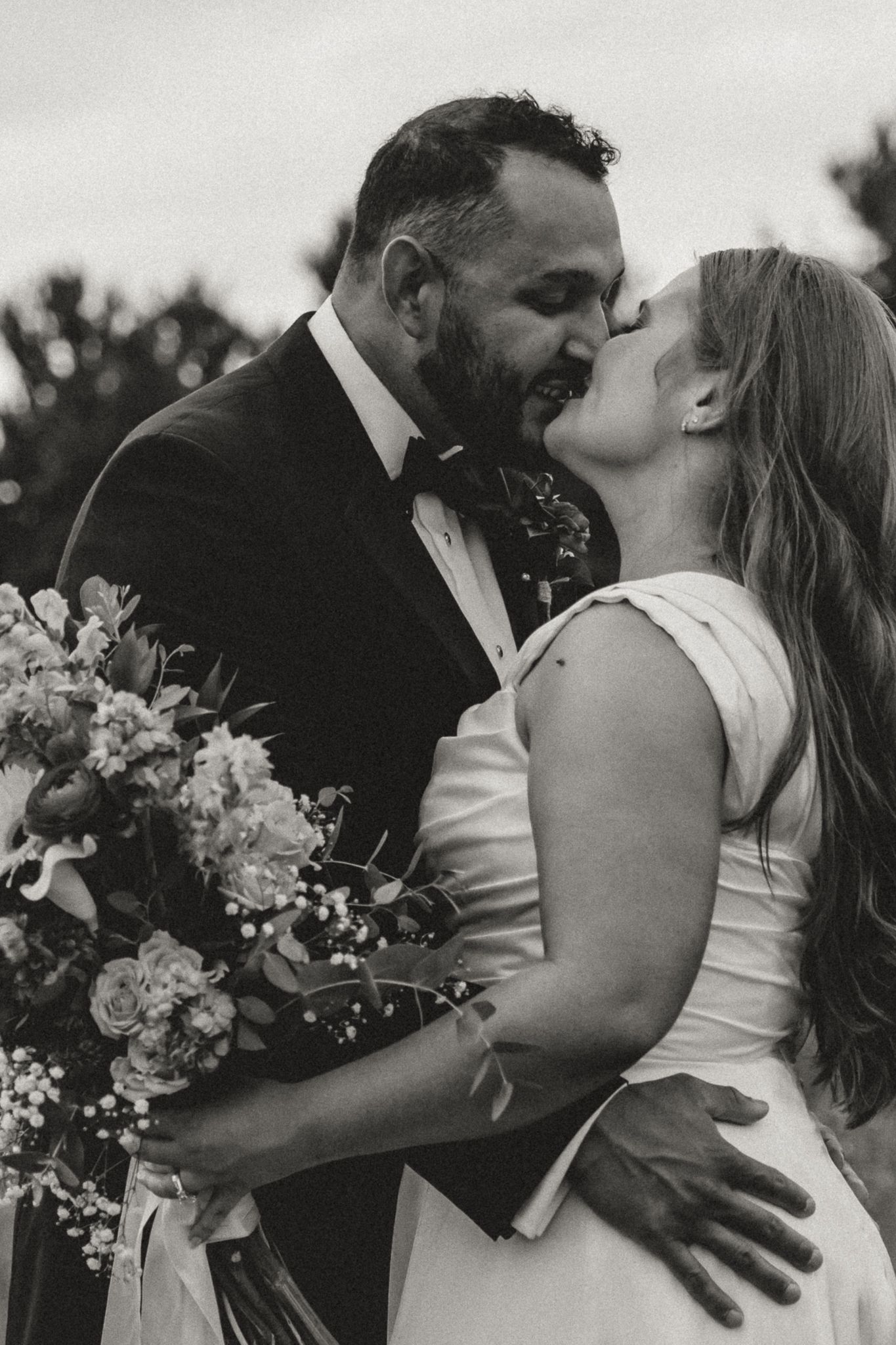 Black and white photo of a bride and groom sharing a kiss, bride holding a floral bouquet at their wedding.
