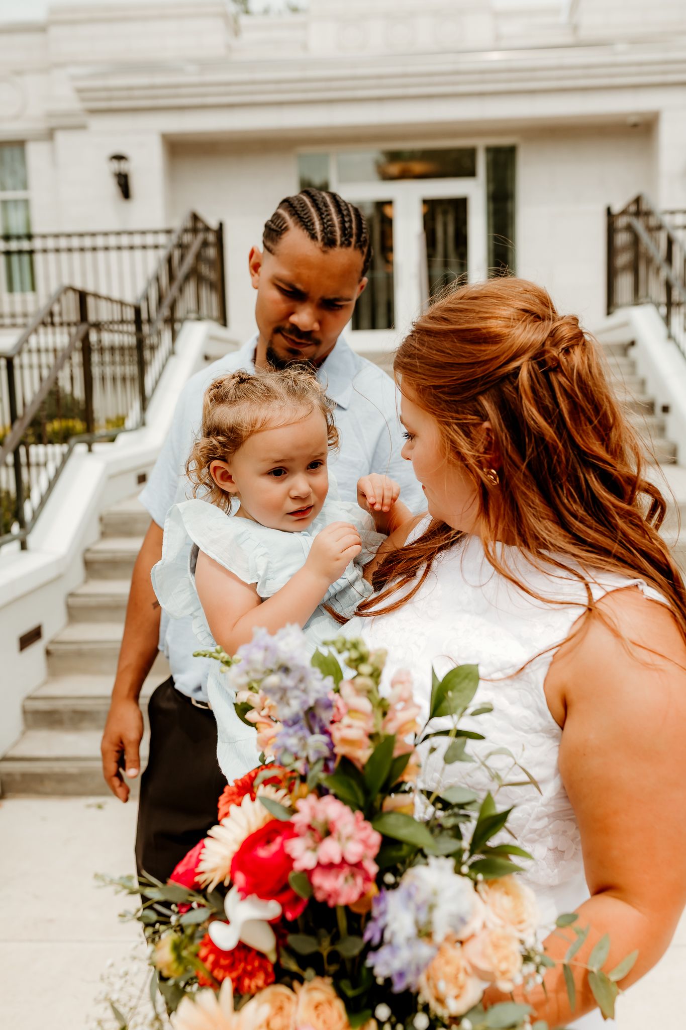 Bride holding colorful bouquet with groom carrying toddler on wedding day outside white building.