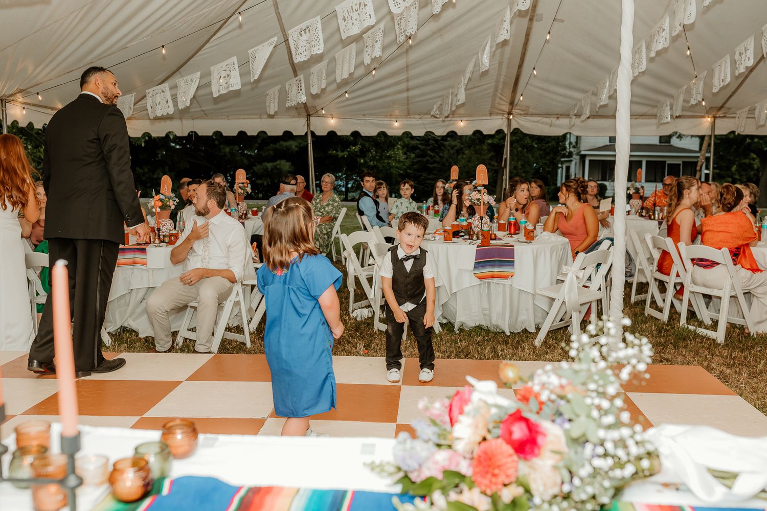 Outdoor wedding reception under a white tent with guests seated at colorful tables, children dancing on a checkered dance floor.