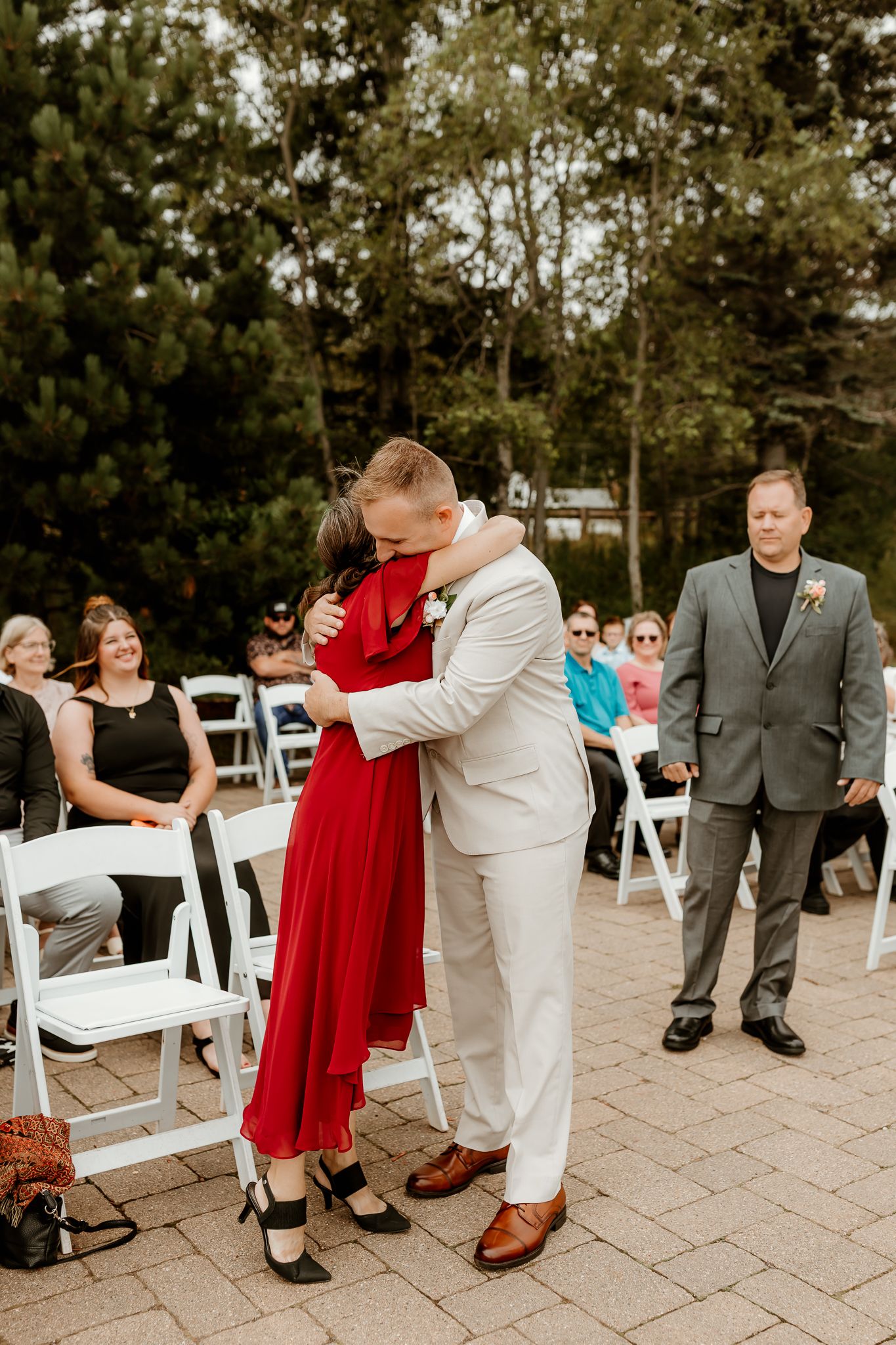 Groom in white suit hugs woman in red dress at outdoor wedding ceremony with seated guests and trees in background.