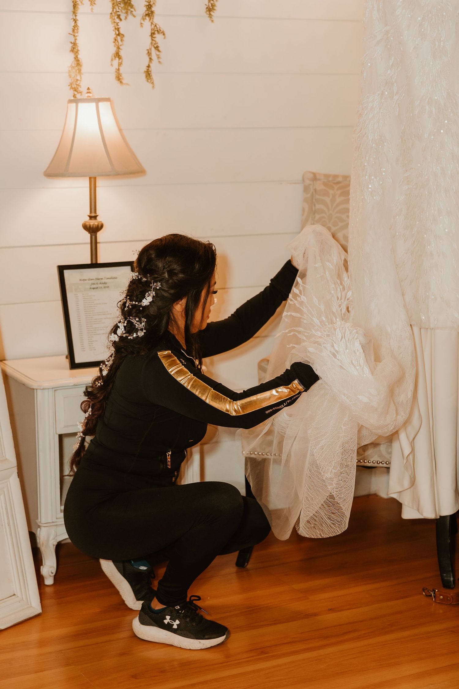 Woman in black tracksuit crouching to arrange a white lace wedding dress hanging in a warmly lit bridal suite.