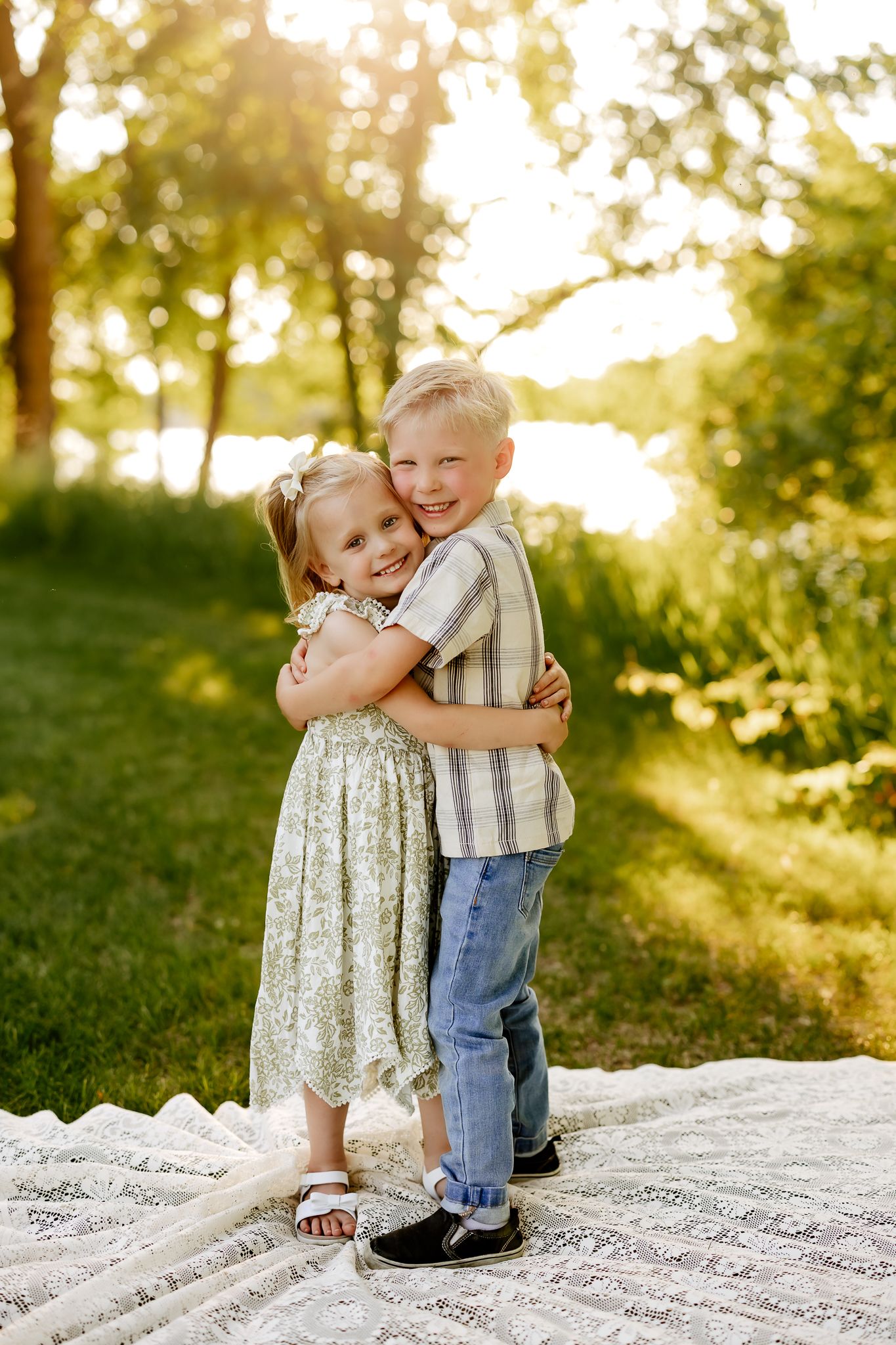 Two young siblings hug and smile on a blanket in a sunny park near a lake.