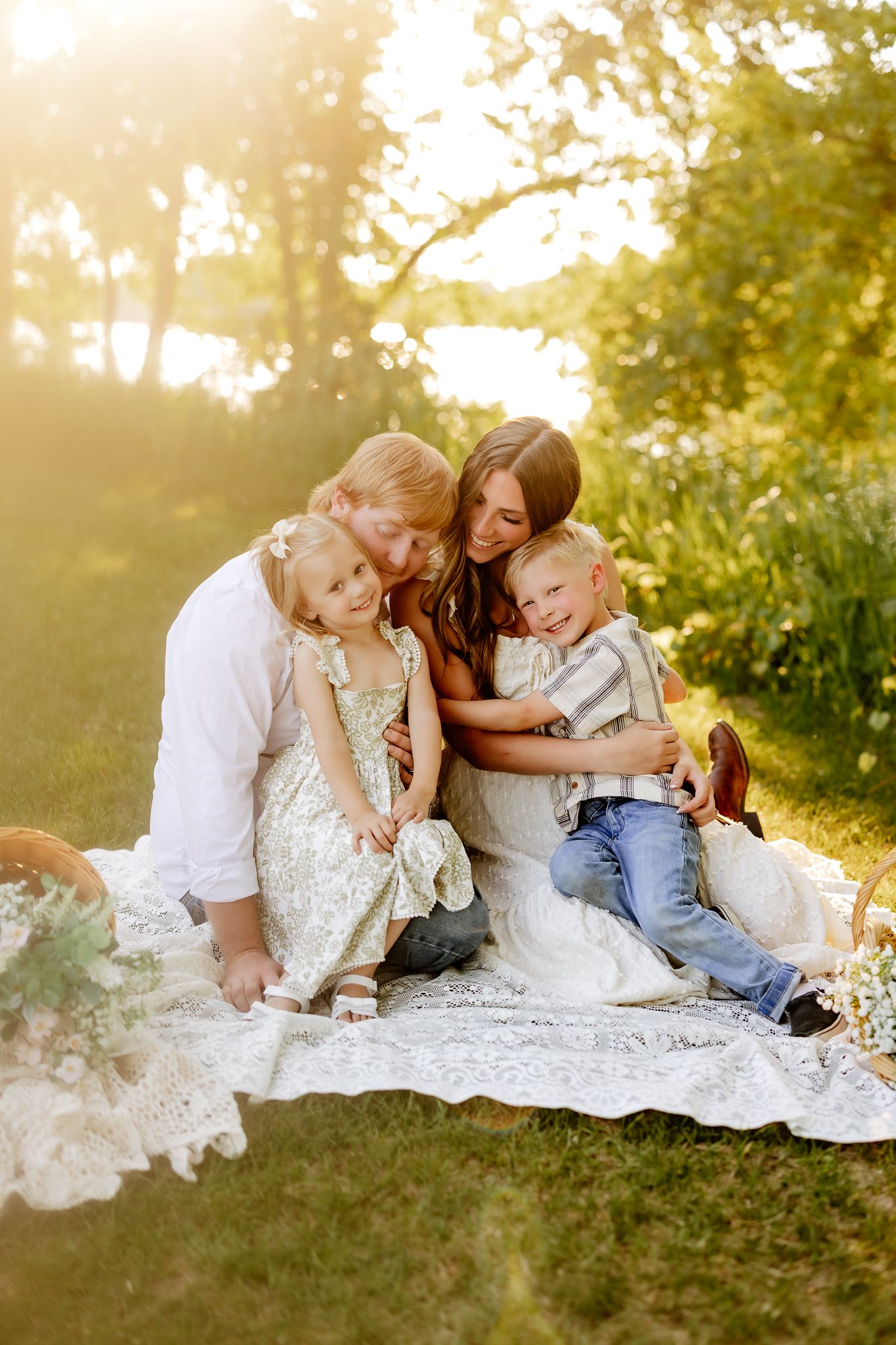 Family of four sitting on a lace blanket in golden sunlight, parents hugging young children outdoors.