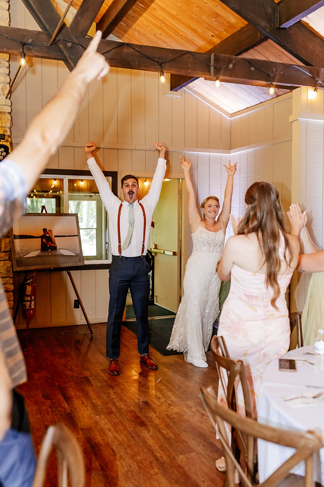 Bride and groom celebrate with raised arms at rustic wedding reception venue with wood ceiling and string lights.