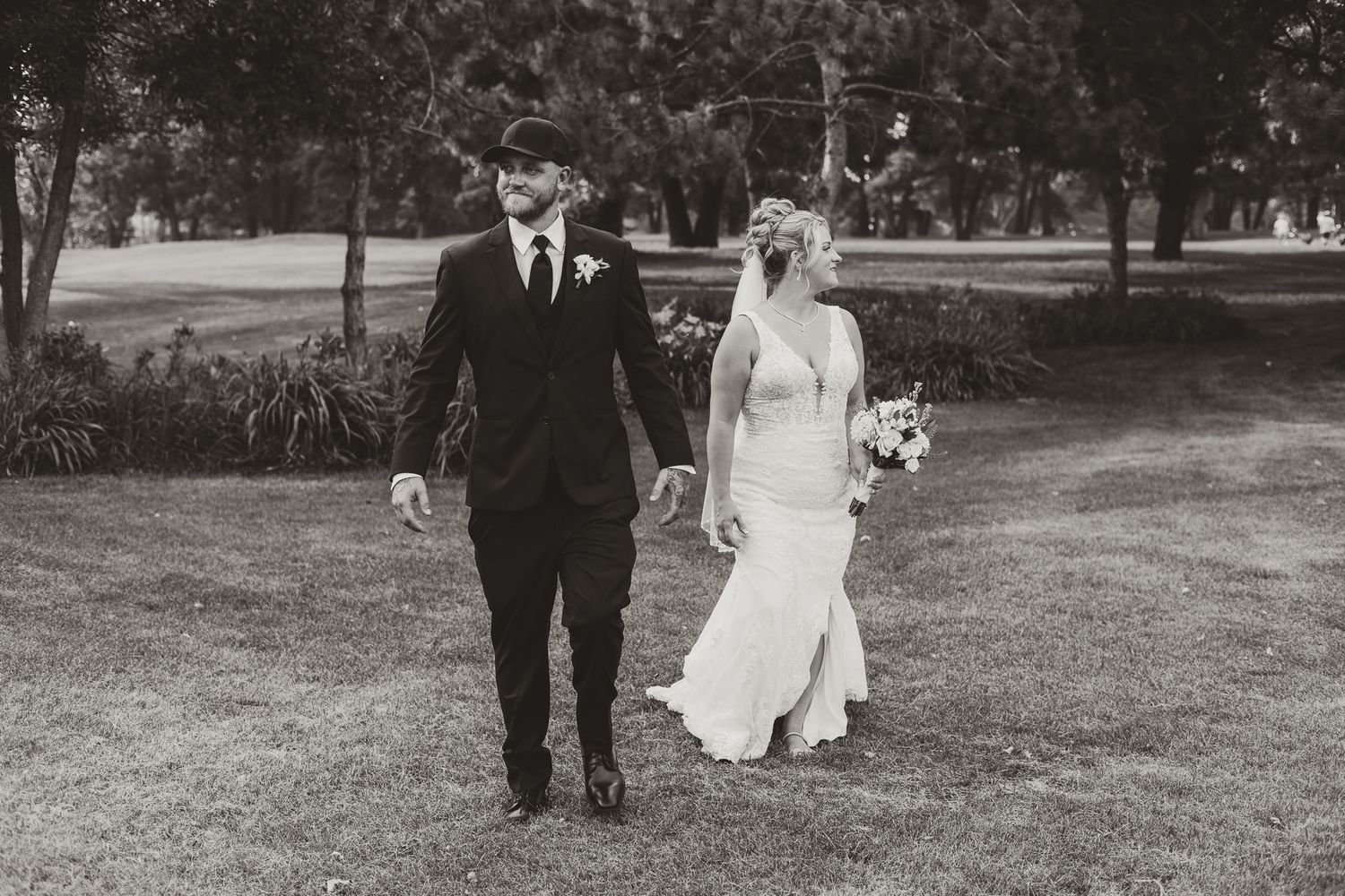 Black and white photo of a bride and groom holding hands walking through a park on their wedding day.