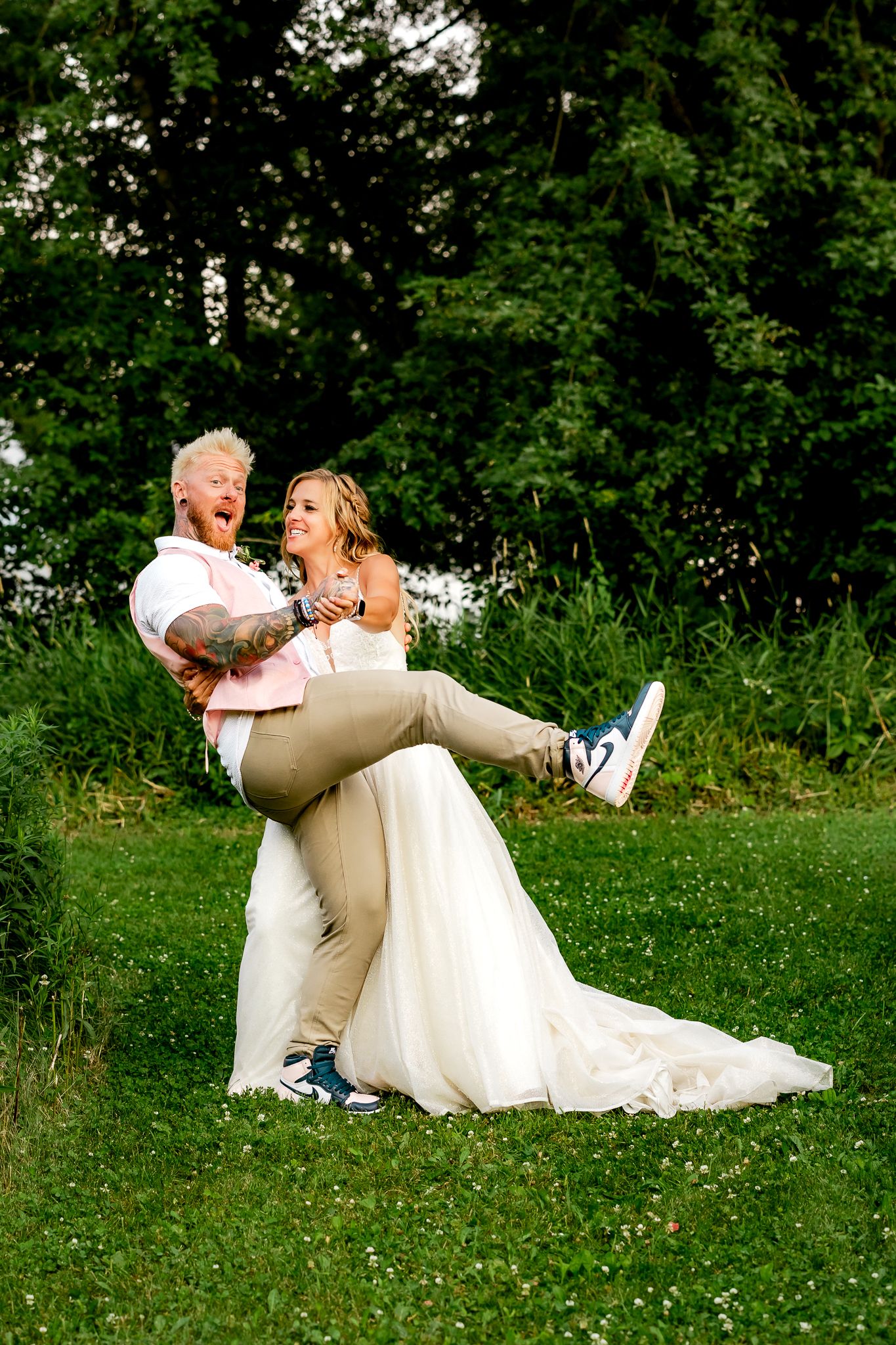 Bride in white gown and sneakers lifted by groom in pink shirt, laughing outdoors on green lawn.