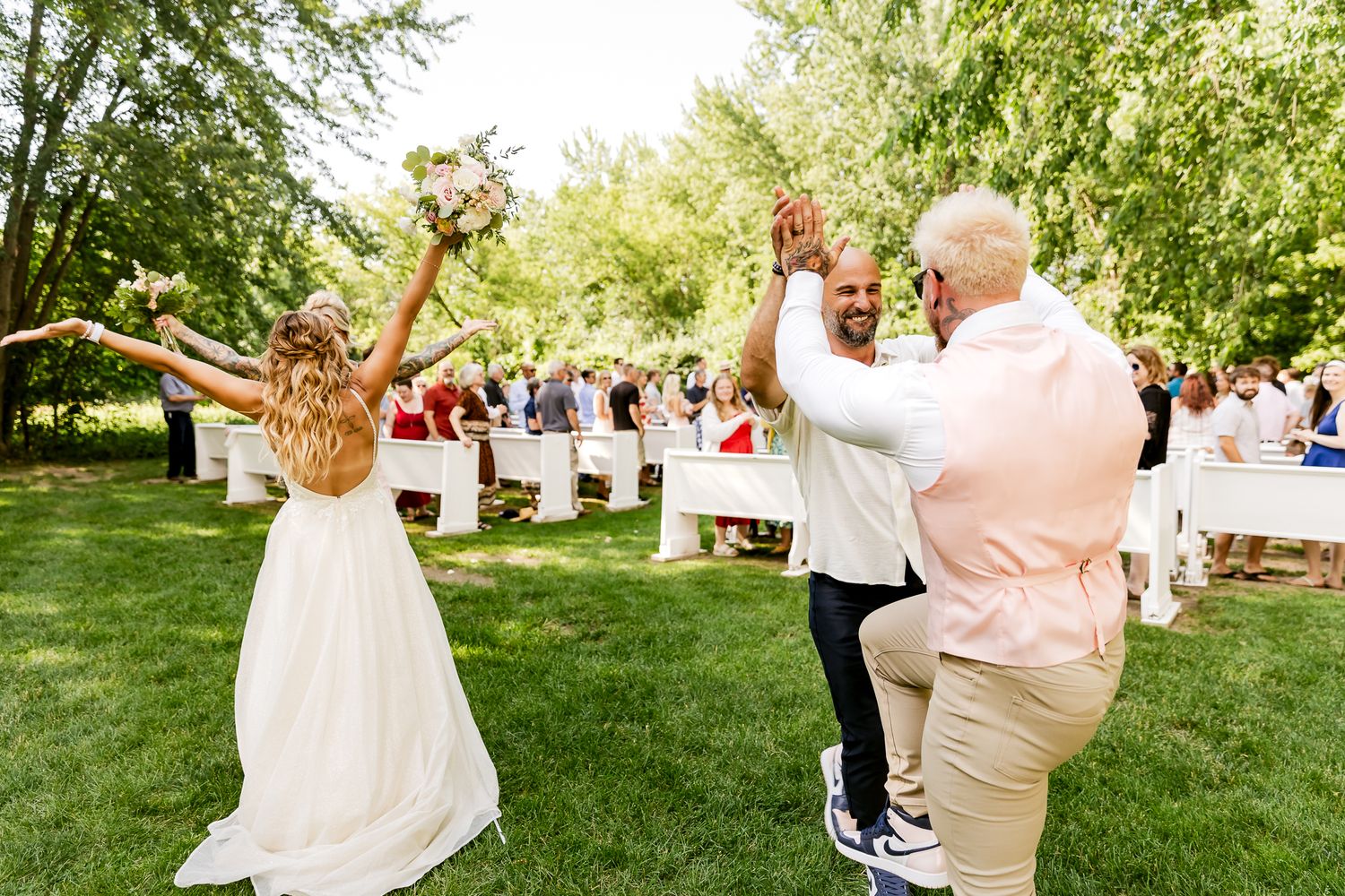 Bride in white gown celebrates with groom and guest at outdoor wedding ceremony surrounded by lush green trees.