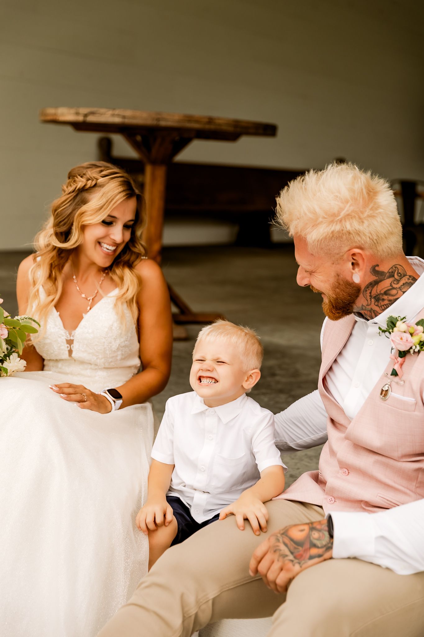 Bride and groom in pink suit sit with young boy in white shirt, laughing together at a rustic wedding venue.