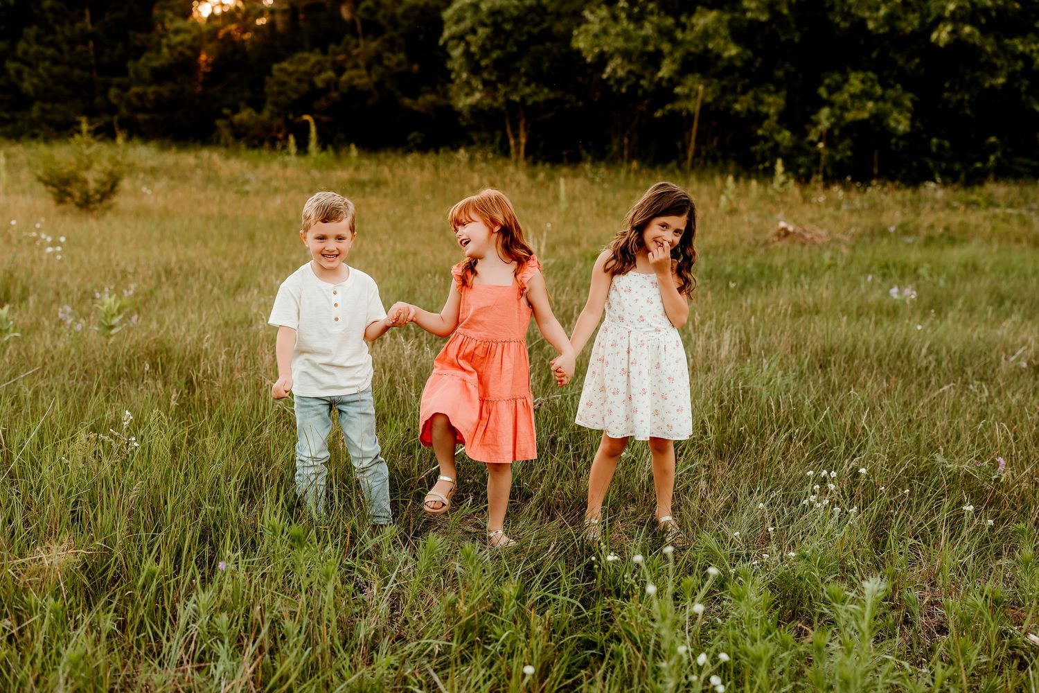 Three children hold hands walking through a sunny green meadow, wearing summer outfits in white, coral, and floral.