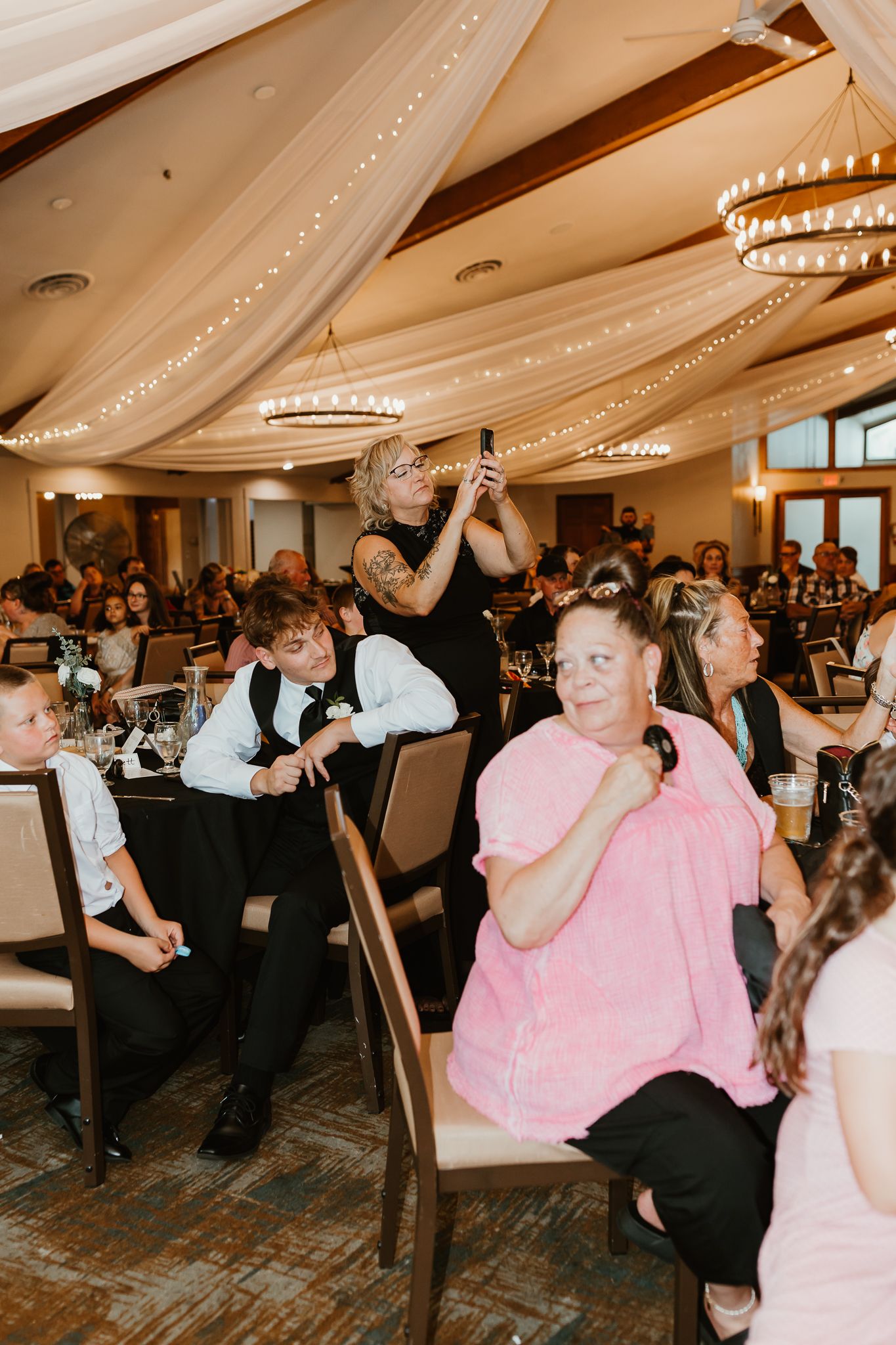Guests seated at a elegant wedding reception venue with draped ceiling lights and chandeliers.