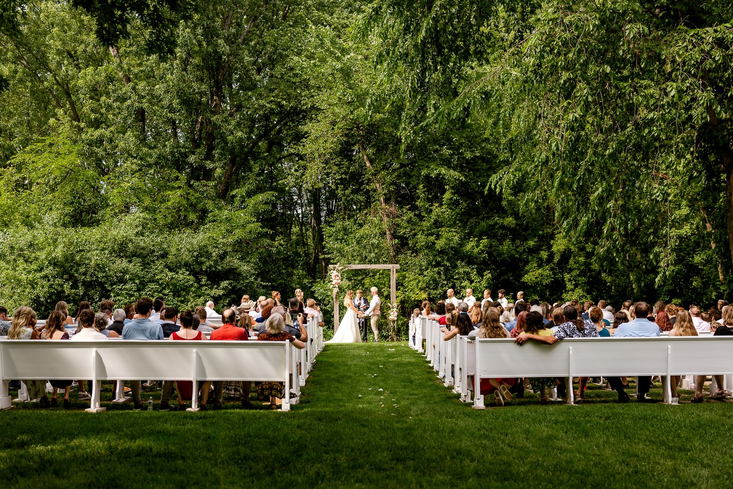 Outdoor wedding ceremony on green lawn with white benches, guests seated, couple at floral arch, surrounded by lush trees.
