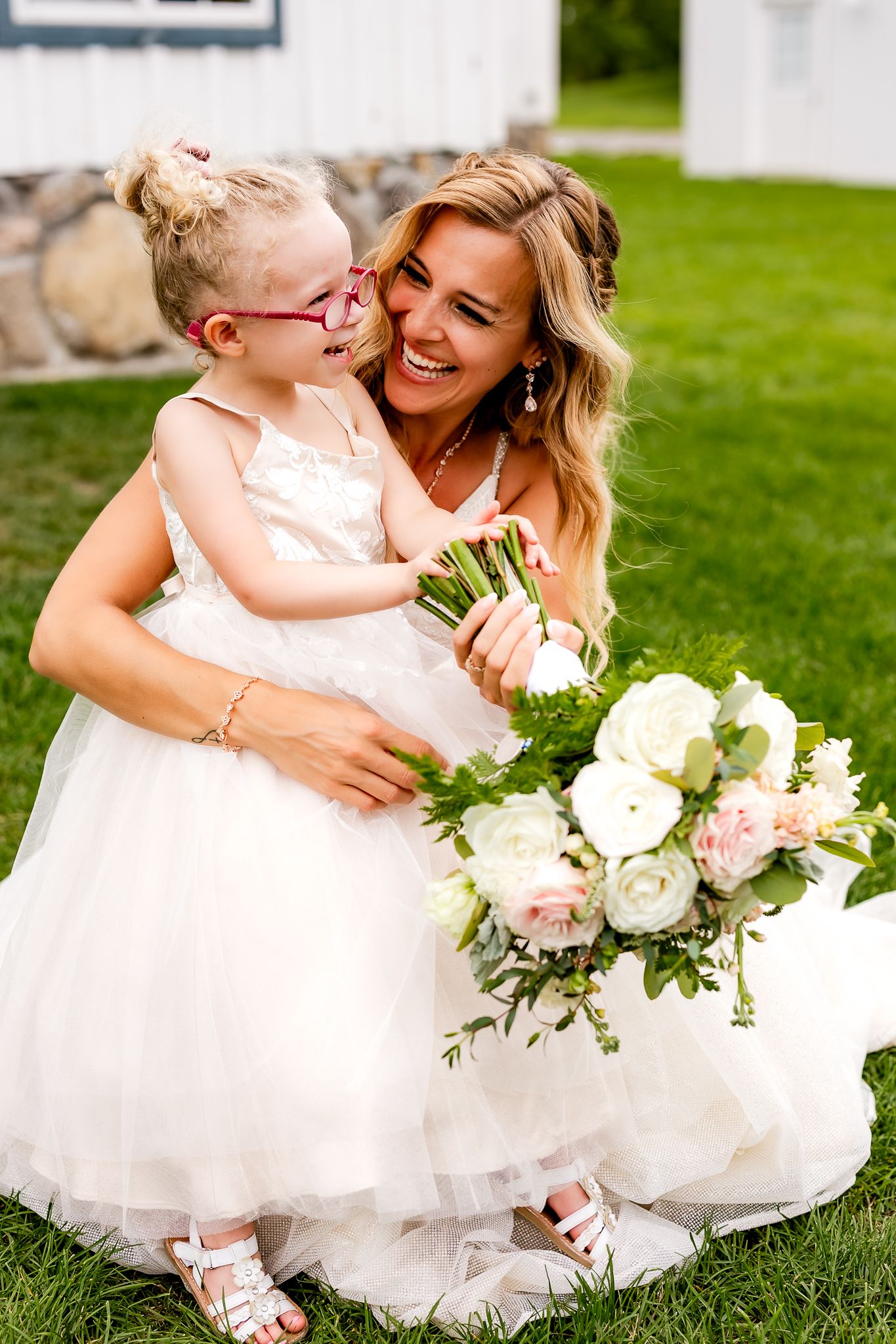 Bride in white gown laughs with flower girl in tulle dress holding bouquet of white and pink roses outdoors.