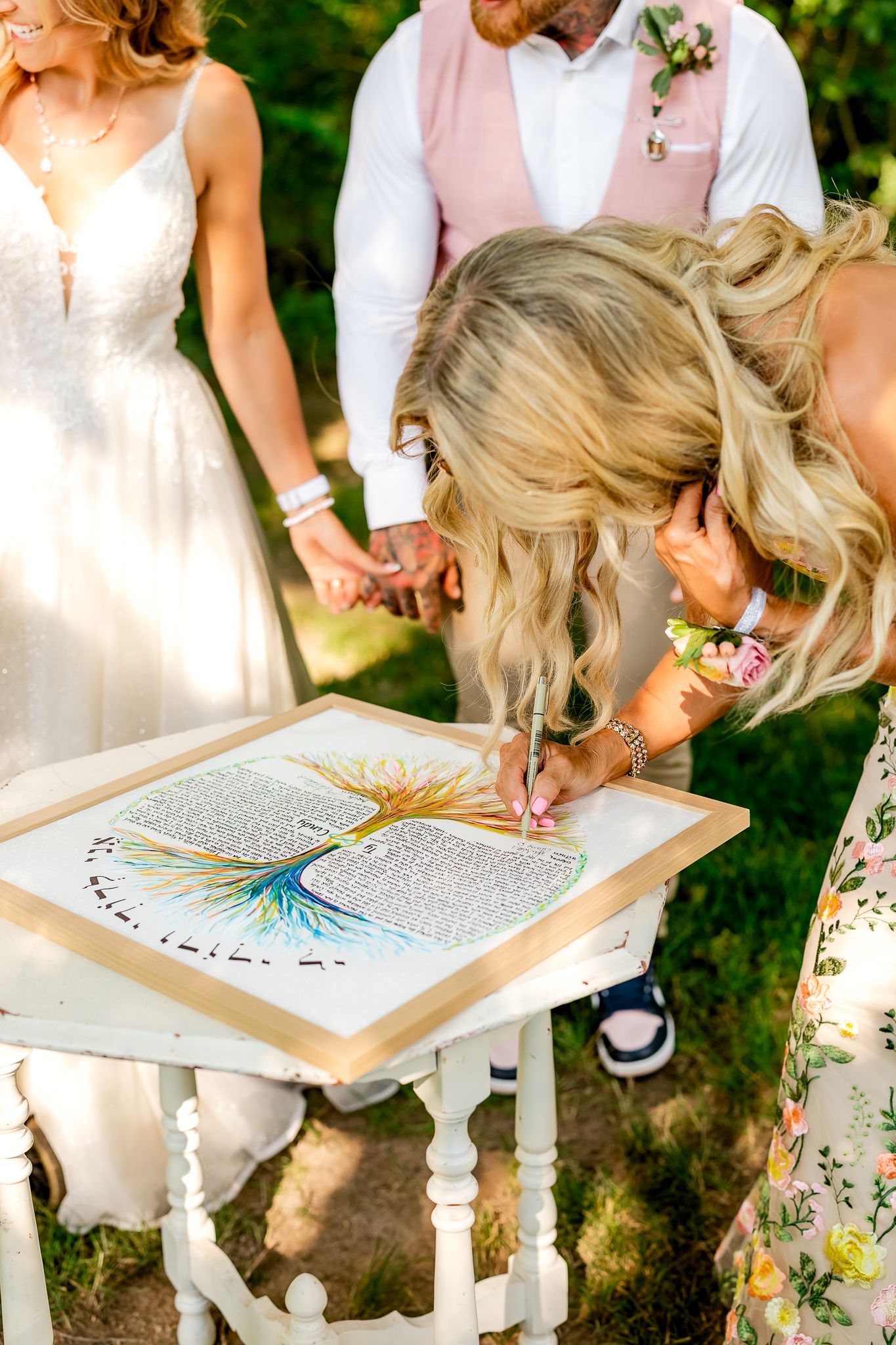 Blonde woman signing a colorful tree ketubah at a wedding ceremony outdoors with bride and groom standing nearby.